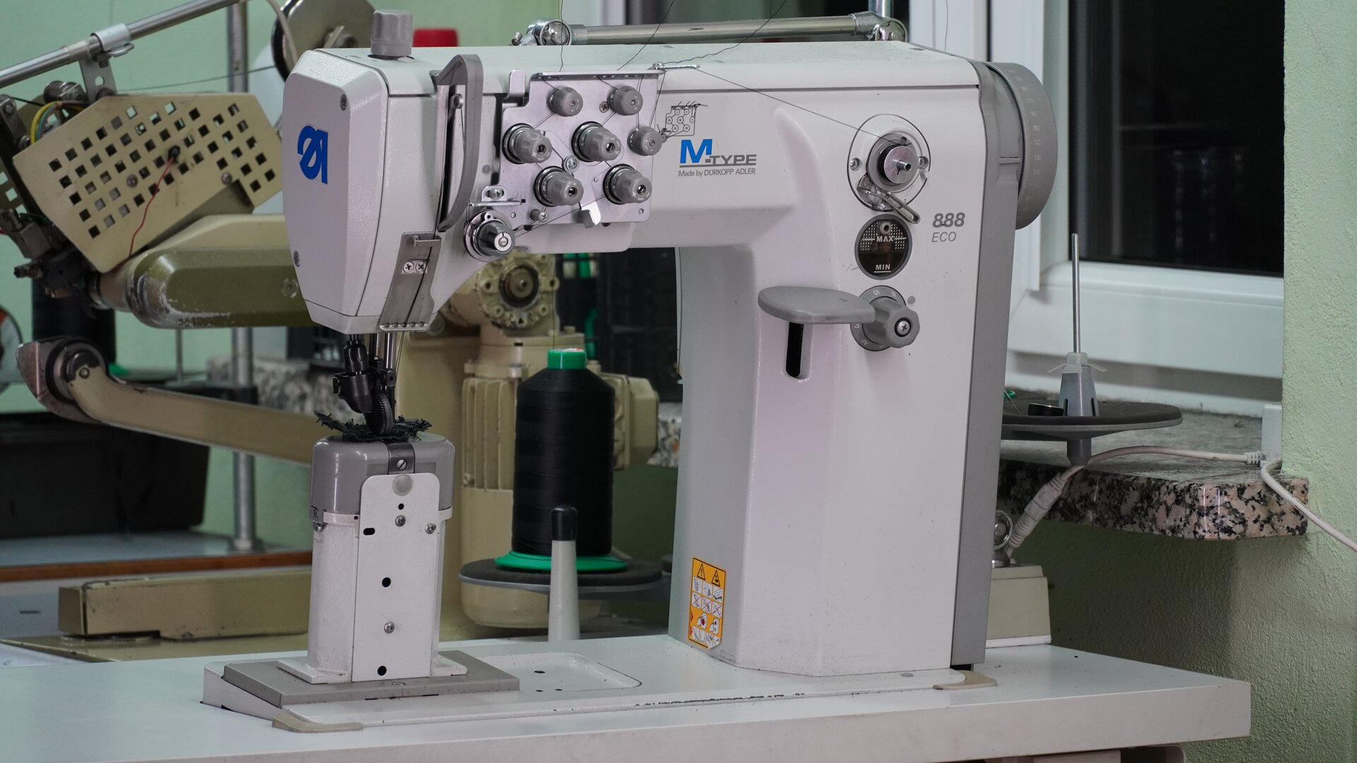 Zdjęcie przedstawia białą maszynę słupkową dwuigłową do szycia, z przodu pośrodku ma siedem pokręteł. Nagranie tożsame z treścią.