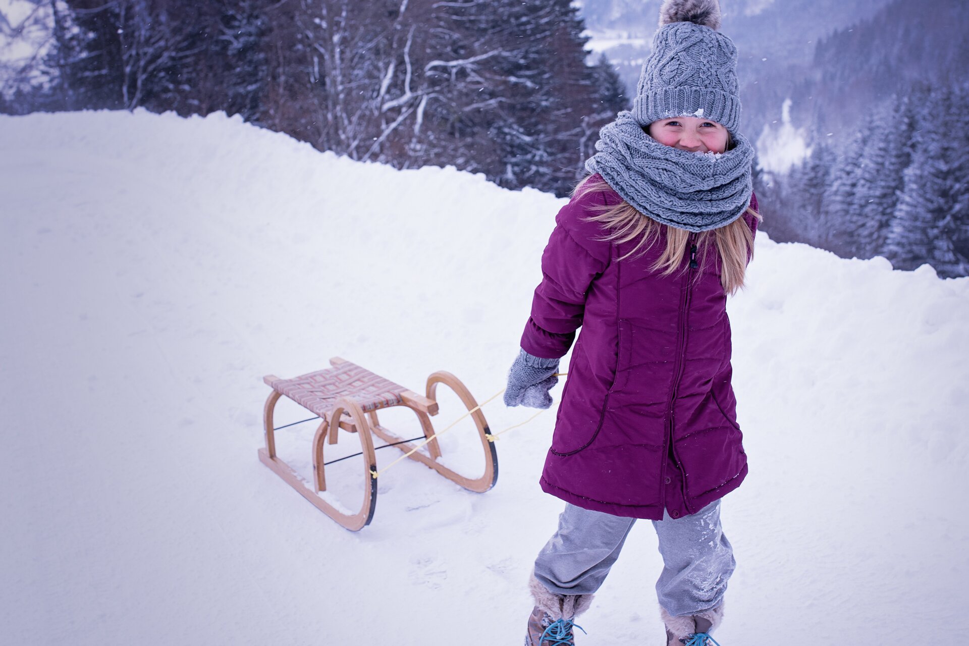 Ilustracja przedstawia ubraną w fioletową kurtkę i szarą czapkę dziewczynkę. Ciągnie ona za sobą drewniane sanki w zimowej scenerii.