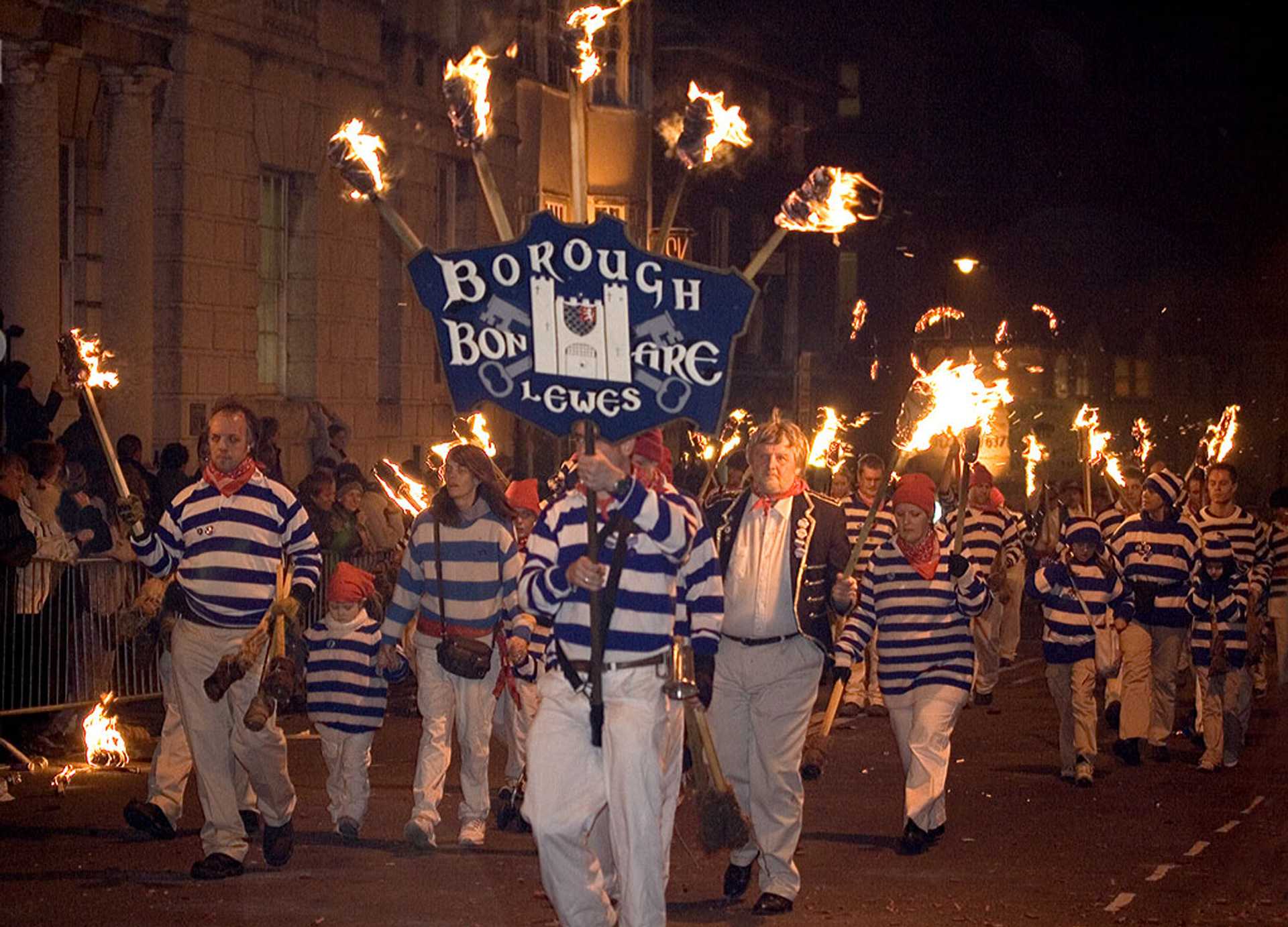 Zdjęcie przedstawia nocną paradę osób, ubranych w jednakowe stroje, czyli białe spodnie i bluzki w biało‑niebieskie paski. Osoby niosą w rękach zapalone pochodnie. Na przodzie widoczna jest postać niosąca konstrukcję z transparentem i pięcioma zapalonymi pochodniami na górze. Transparent, w kolorze granatowym, pośrodku ma schematycznie przedstawioną fasadę zamku, a u góry, po bokach i na dole widoczne są napisy z białych liter w języku angielskim:Borough Bonfire Lewes.Jest to nazwa stowarzyszenia z miejscowości Lewes. W tle, po lewej stronie, widoczne fasady budynków.