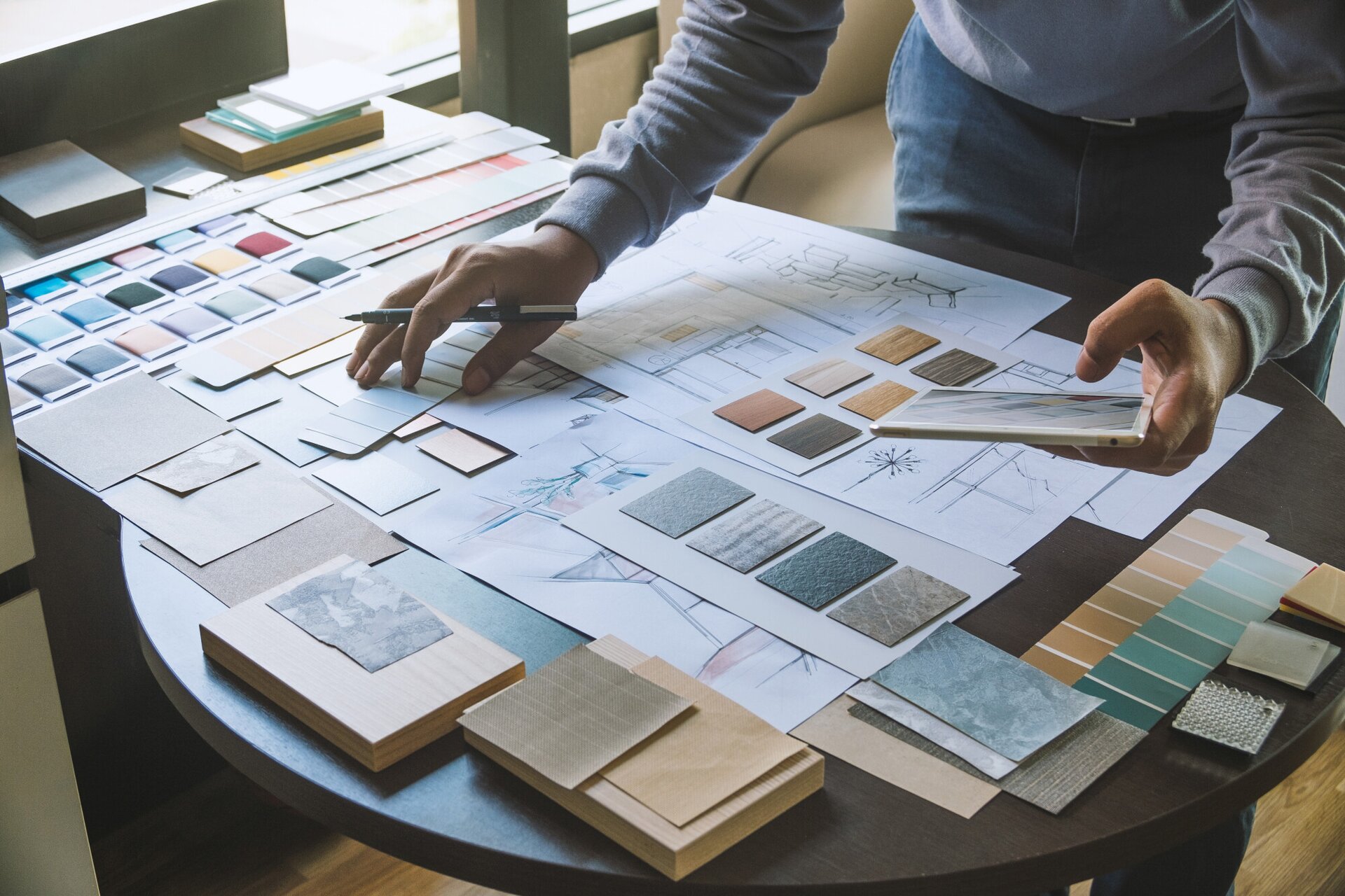 Zdjęcie przedstawia projektanta wnętrz podczas pracy. Mężczyzna ma na sobie niebieskie jeansy oraz jasną, szarą bluzę z długim rękawem. Znajduje się w pomieszczeniu, stoi przy brązowym, drewnianym, okrągłym stole, na którym znajduje się mnóstwo próbek materiałów. Projektant w lewej dłoni trzyma tablet, a w prawej długopis.