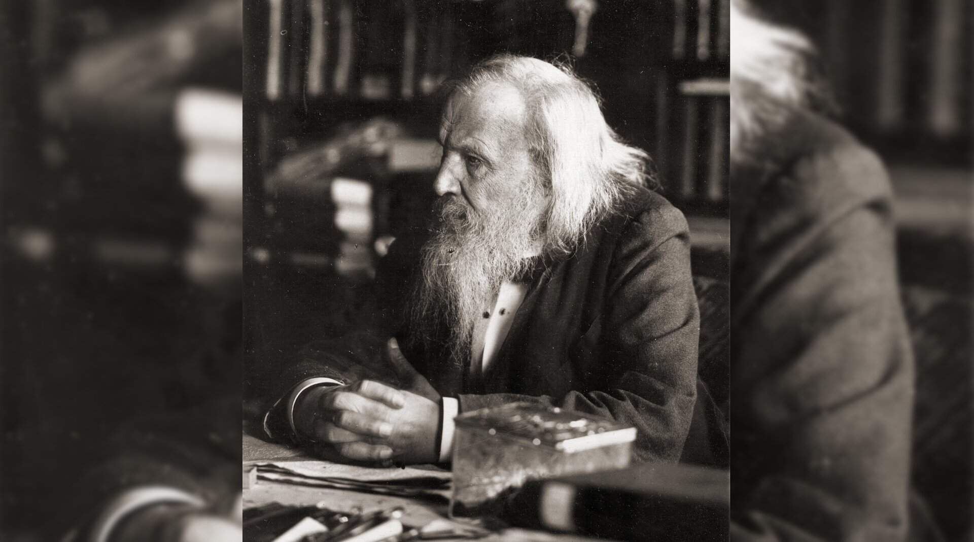 Fotografia przedstawiająca starszego mężczyznę z siwą brodą i długimi włosami, siedzącego przy biurku.