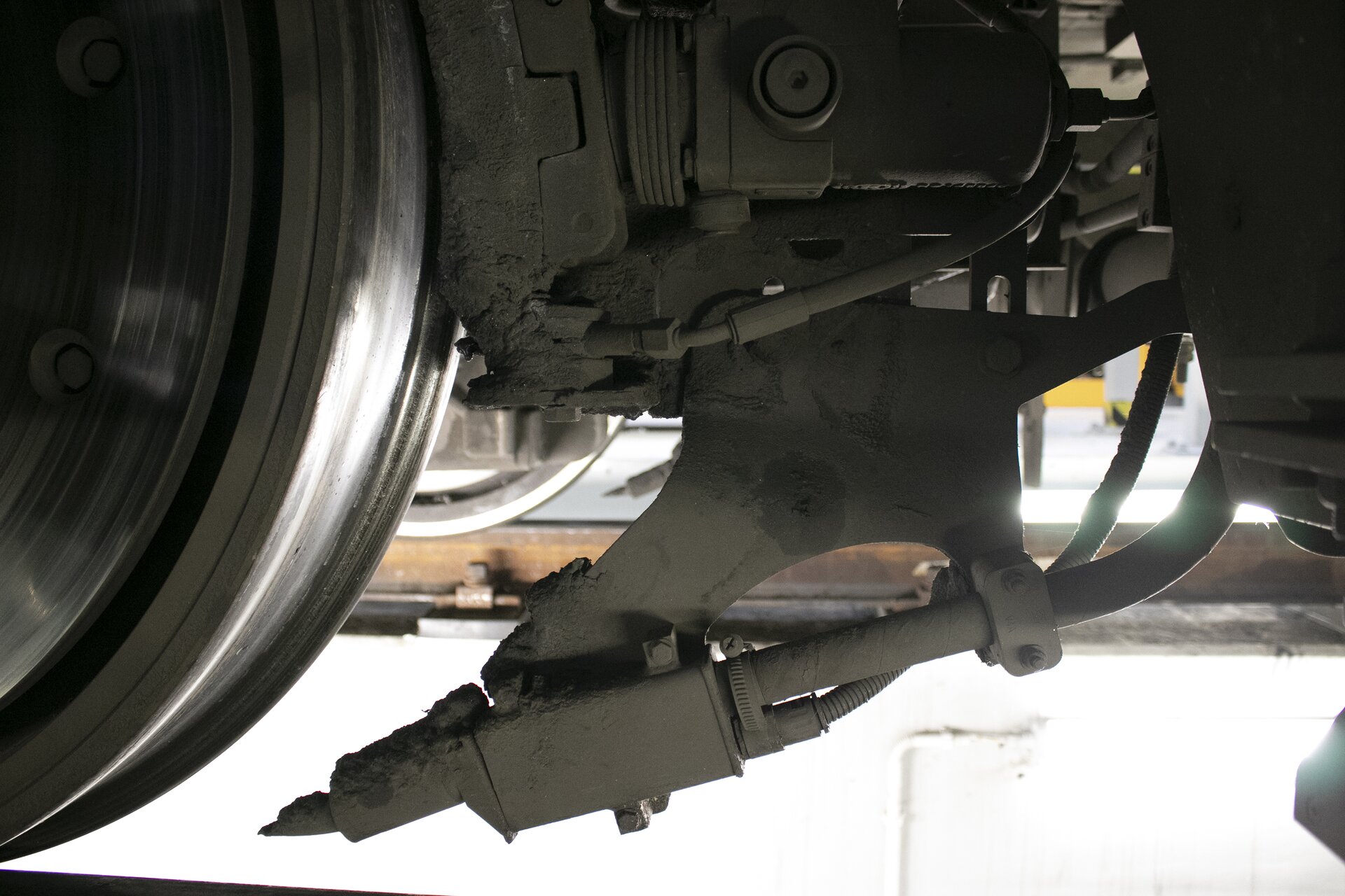 Zdjęcie przedstawia piaseczniczę. Zdjęcie zrobione zostało spod wagonu pociągu. Z lewej strony zdjęcia widać fragment koła. Na tym zdjęciu piasecznica widoczna jest wyraźnie, ustawiona tuż przed kołem. Dysza piasecznicy skierowana jest w lewo, w miejsce styku koła z szyną. Piasecznica znajduje się obok kół pociągu, wyglądem przypomina rurę wydechową. Służy ona do posypywania szyn piaskiem w celu zwiększenia tarcia pomiędzy kołem a szyną w celu przywrócenia utraconej przez koło przyczepności lub w celu zapobiegnięcia utracie przyczepności.