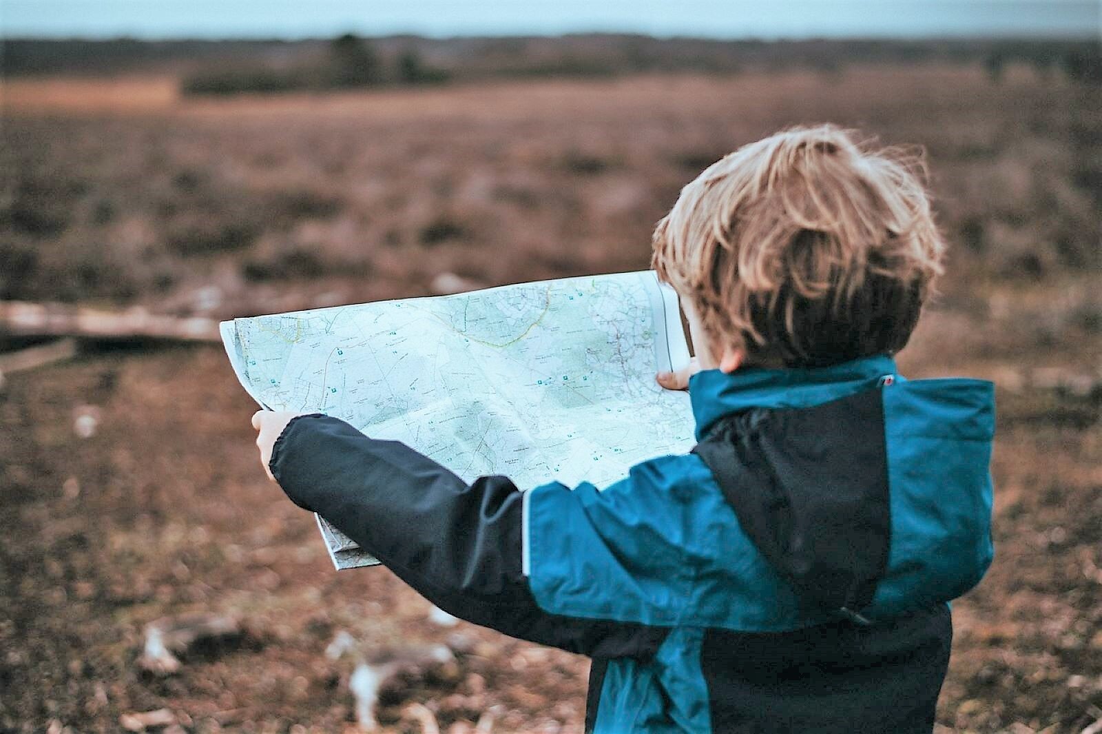 Zdjęcie ukazuje odwróconego tyłem chłopca w kurtce, z rozłożoną mapą trzymaną w rękach. Przed nim rozpościera się rozległa przestrzeń.