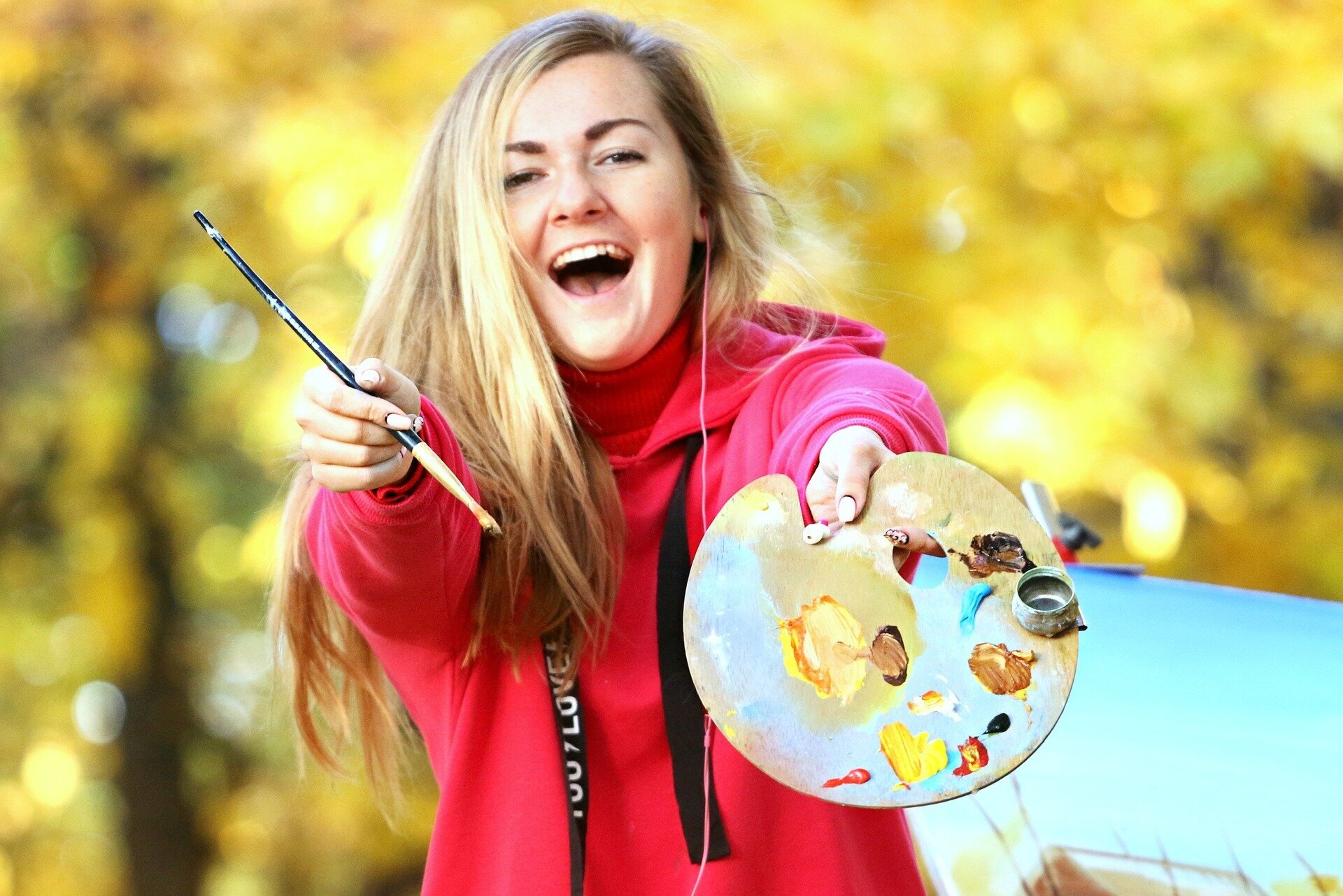 Zdjęcie przedstawia młodą, długowłosą kobietę. Kobieta śmieje się, ma szeroko otwarte usta. W dłoniach trzyma paletę do malowania z różnokolorowymi farbami oraz pędzel. Za jej plecami, w tle drzewa z żółtymi liśćmi. 