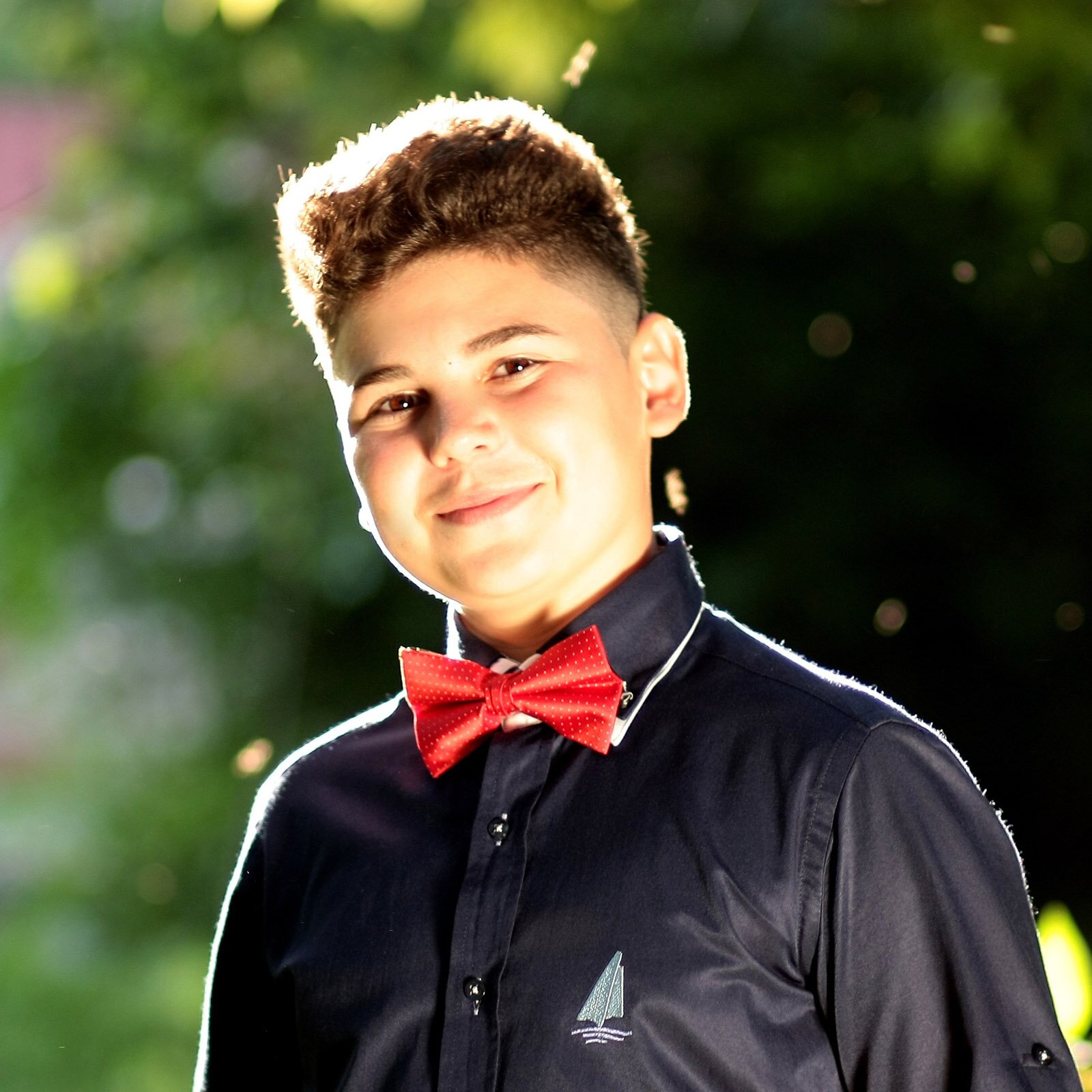 Zdjęcie przedstawia uśmiechniętego młodego chłopaka, w ujęciu od klatki piersiowej w górę, ubranego w elegancką, czarną koszulę z malutkim nadrukiem żaglówki po lewej stronie oraz czerwoną muszkę. Chłopiec ma pogodny wyraz twarzy i lekko się uśmiecha.