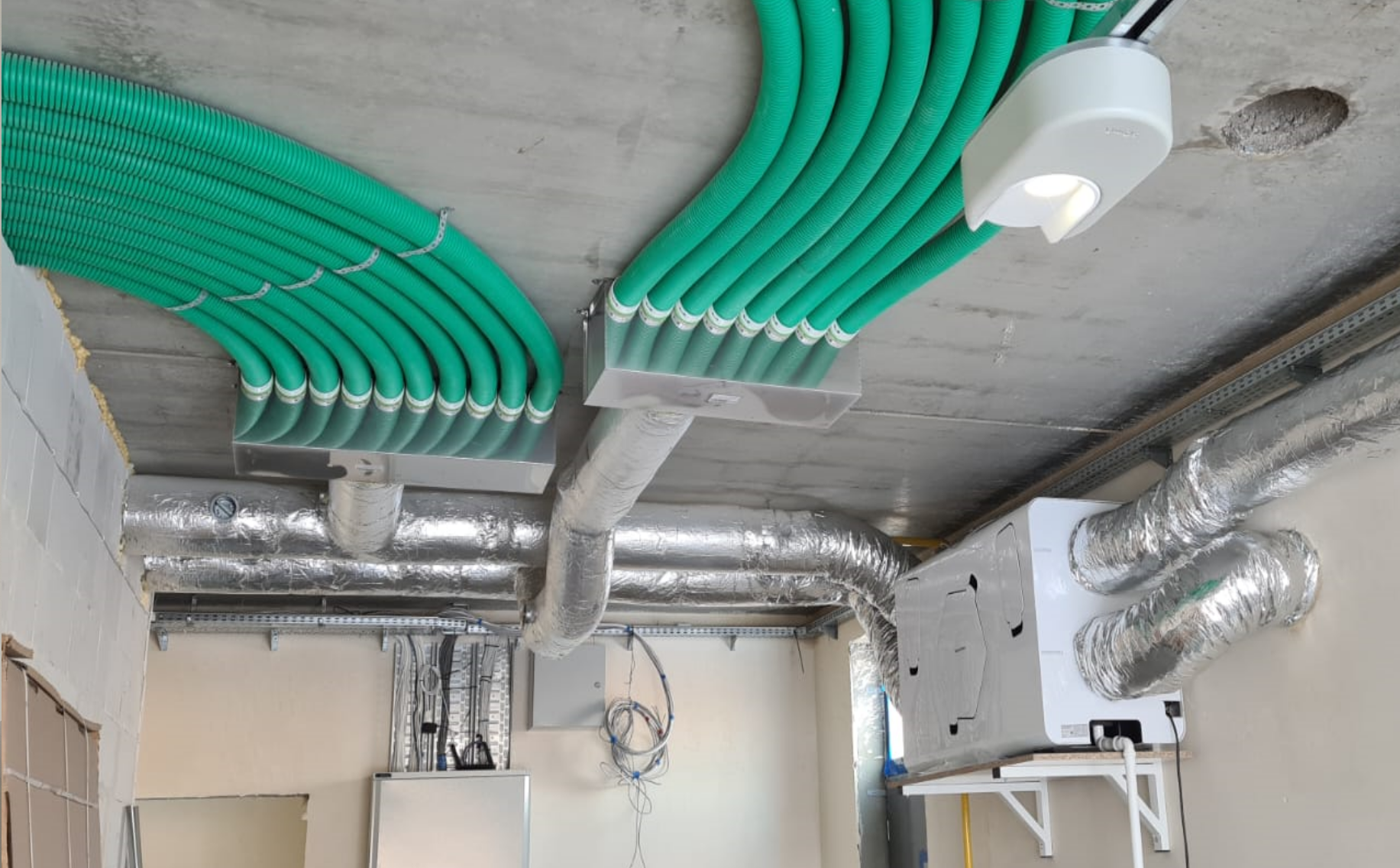 Zdjęcie przedstawia centralę wentylacyjną z odzyskiem ciepła wraz z izolowanymi termicznie kanałami. Widoczne są rzędy rur podwieszone pod sufitem, które łączą się w izolowanych termicznie kanałach.