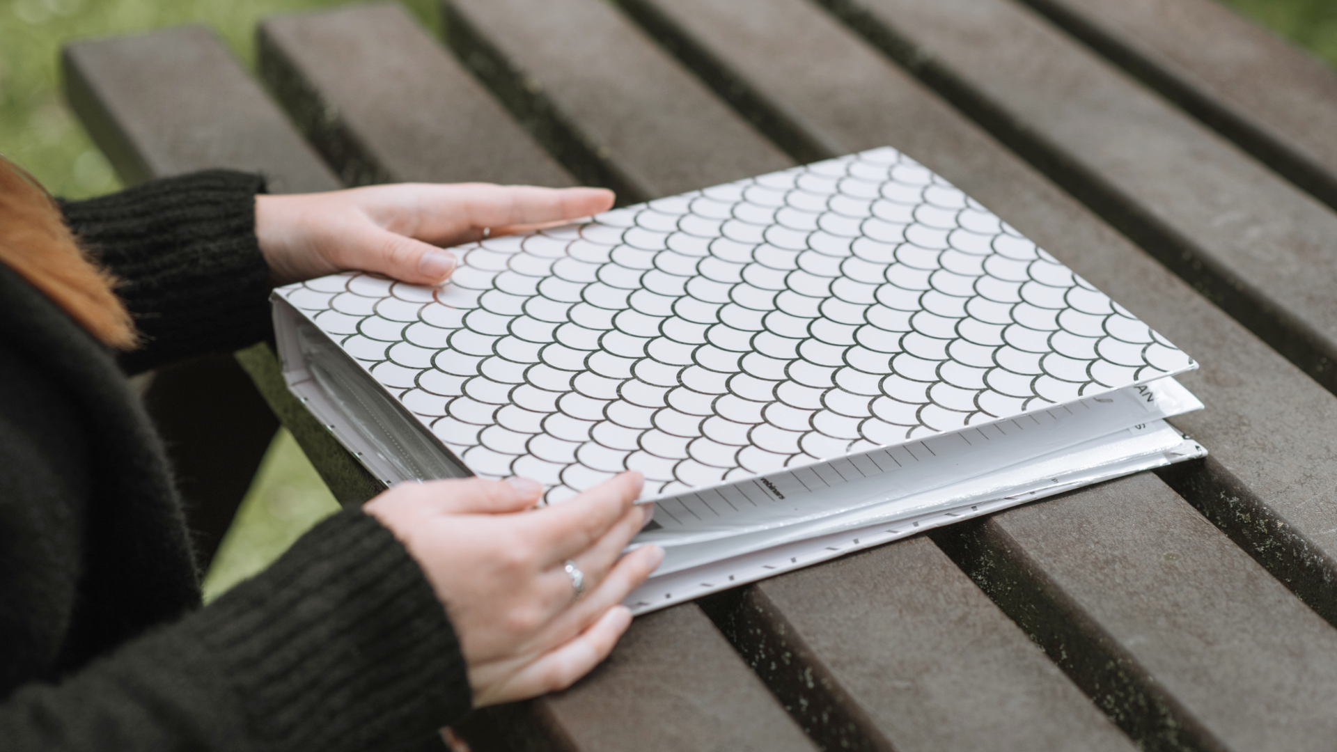 Zdjęcie przedstawia teczkę, która leży na stole ogrodowym. Przy stole znajduje jest dziewczyna/kobieta, której widoczne są tylko dłonie. Kobieta trzyma dłonie na teczce.