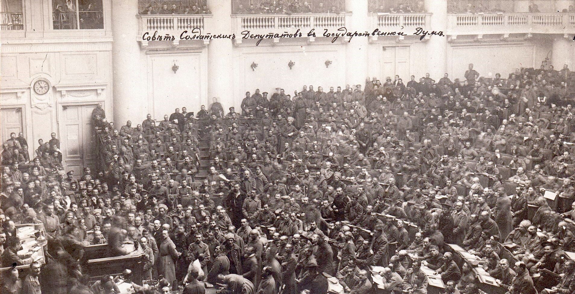Zdjęcie przedstawia ogromną salę z mnóstwem osób. W sali są pulpity i siedzenia ułożone rzędami. Siedzą tam ludzie. Część osób stoi. Większość zgromadzonych ma na sobie płaszcze i mundury wojskowe. 