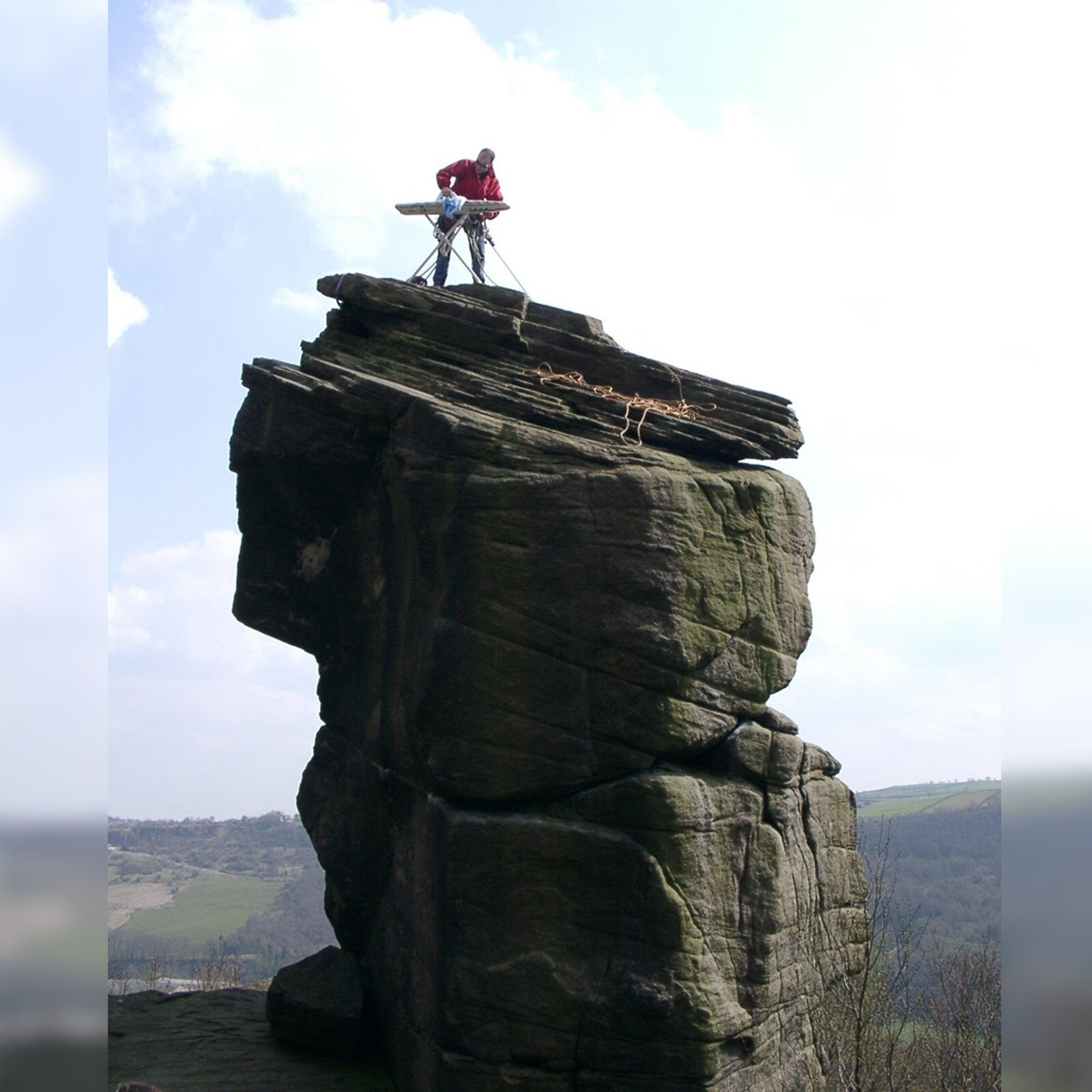 Zdjęcie przedstawia mężczyznę stojącego na szczycie samotnej góry. Widoczna jest deska do prasowania, na której mężczyzna prasuje. 