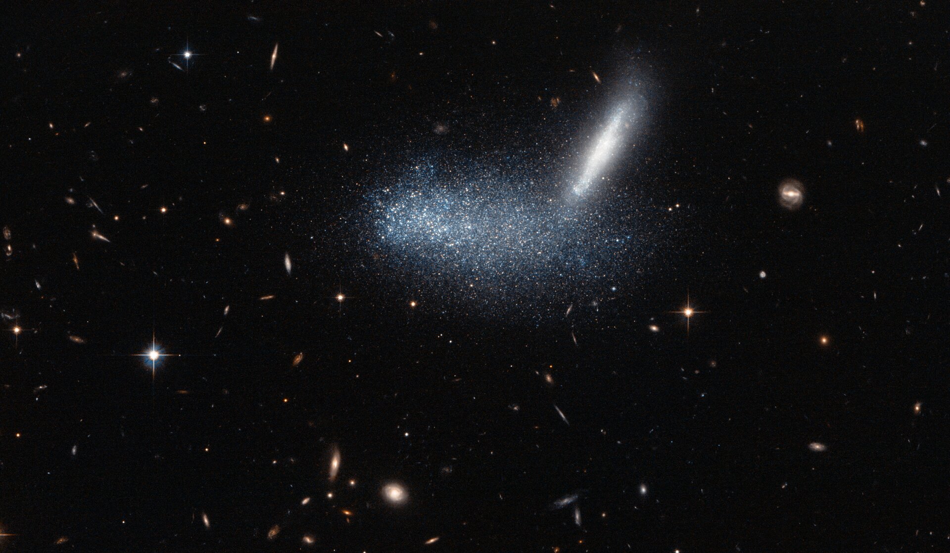 Grafika przedstawia nieregularną galaktykę karłowatą PGC 16389, zakrywającą sąsiednią galaktykę APMBGC 252 + 125-117. Na zdjęciu znajduje się skupisko jasnych punktów. Na górze z jego prawej strony jest jasne, pionowe pasmo. Wokół na czarnym tle świecą jasne punkty.  