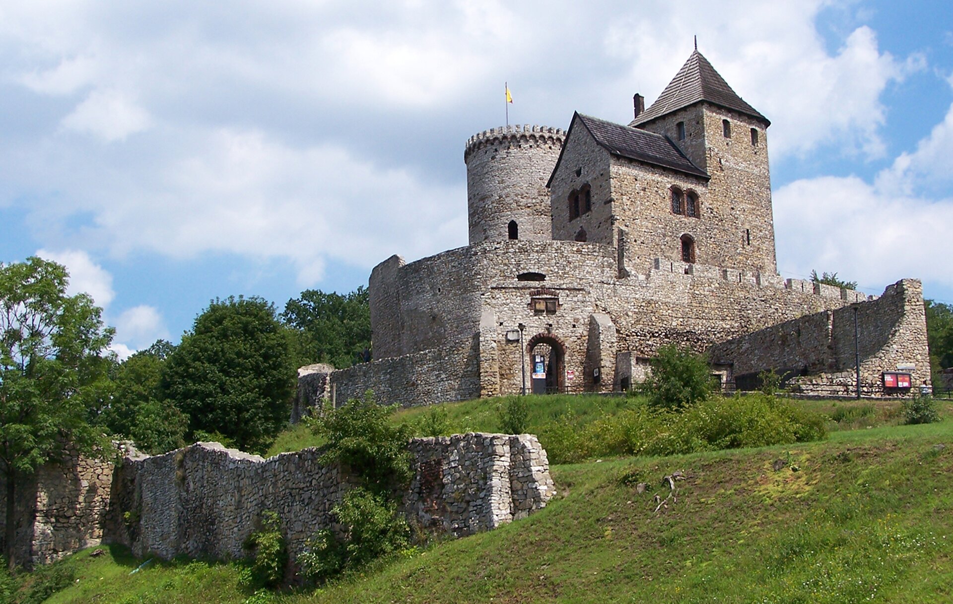 Zdjęcie przedstawia zamek stojący na porośniętym zieloną trawą wzgórzu. Okala go mur. Za nim znajdują się zabudowania, zamku z wieżą i basztą. U stóp wzgórza są pozostałości drugiego muru.