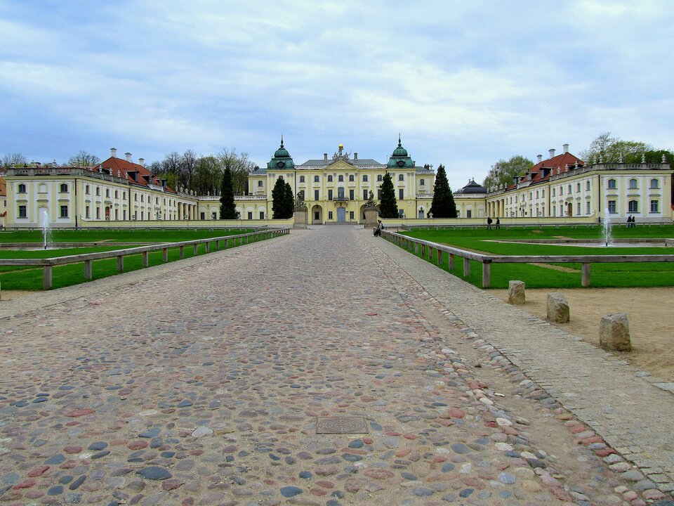 Ilustracja przedstawia Pałac Branickich, Białymstoku. Ilustracja ukazuje pałac wraz z brukową drogą prowadzącą do niego oraz fragment ogrodu. Widoczny jest duży, jasny budynek o układzie horyzontalnym i dużej liczbie okien rozmieszczonych w stałych odstępach. Bryła budynku ma kształt litery C. Część środkowa jest trójkondygnacyjna, boczne - dwukondygnacyjne.  Budynek jest dekorowany rzeźbami.