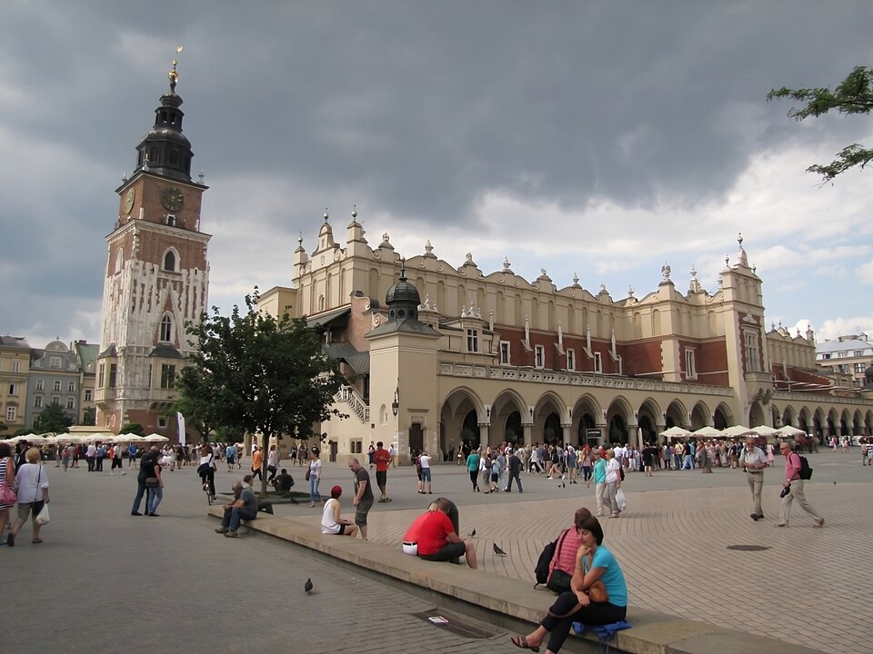 Zdjęcie przedstawia współczesny widok na krakowskie Sukiennice i wieżę dawnego ratusza miejskiego. Renesansowa zabudowa sukiennic i widocznej jej ściany frontowej od strony Kościoła Mariackiego wyróżnia się arkadami na parterze o pełnych łukach otworów i bogato zdobioną attyką u szczytu. Pomiędzy arkadami a attyką długa wnęka ściany z otworami okiennymi i wykusz pośrodku z centralnym wejściem do wnętrza obiektu. Skrzydło wschodnie częściowo zasłonięte przez drzewo na rynku, widać jednak jedną z wież narożnych sukiennic z hełmem na szczycie. Bliżej lewej krawędzi fotografii wieża ratuszowa ceglana, uzupełniona kamieniem w połowie wysokości. Wieża zwęża się ku szczytowi. Poniżej kopuły z hełmem widoczne tarcze zegarowe na dwóch ścianach pod szczytem. U dołu wieży ratuszowej wykusze z oknami. Na pierwszym planie fotografii widać grupy przechodniów i siedzących na ławach turystów. 