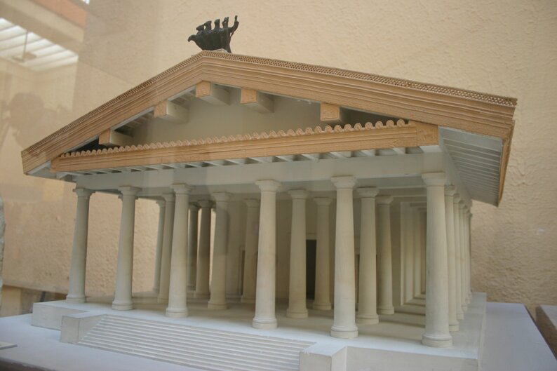 Zdjęcie przedstawia makietę pogańskiej świątyni. Fronton wsparty jest na potrójnej kolumnadzie, a za nim znajduje się niewielki budynek świątyni.