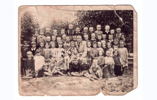 Kliknij, aby powiększyć Czarnobiałe zniszczone zdjęcie przedstawia grupę kilkudziesięciu dzieci w wieku wczesnoszkolnym ustawionych w czterech rzędach na tle krzewów. Między nimi, po środku drugiego rzędu siedzi Bronisław Tokaj - mężczyzna w średnim wieku, w koszuli z krawatem i płaszczu.