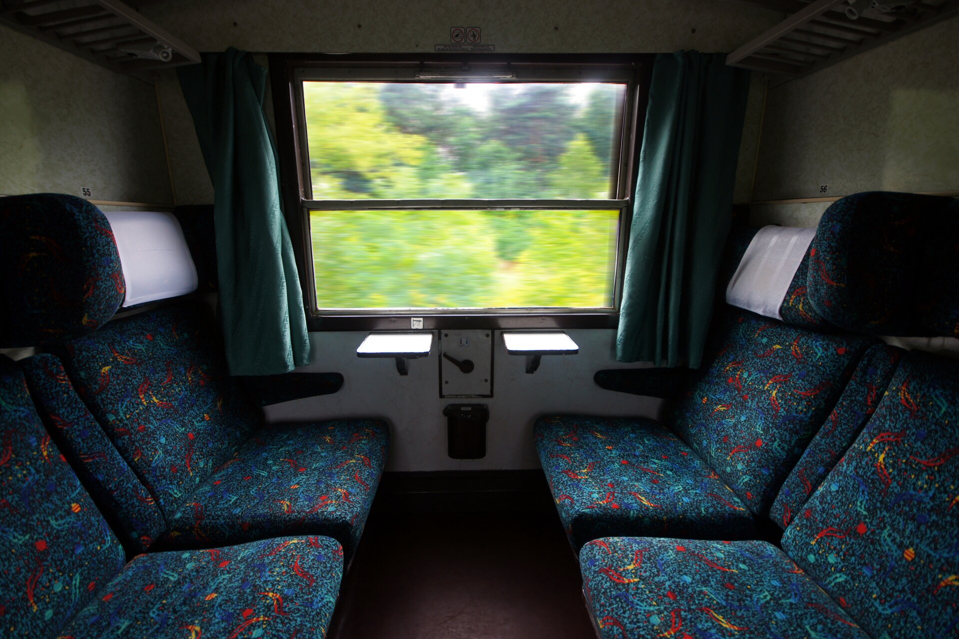 Zdjęcie przedstawia wnętrze przedziału w pociągu. Po lewej i po prawej krzesła obite wielokolorowym materiałem. Na wprost okno. Za oknem widać rozmazane kontury zielonych drzew.