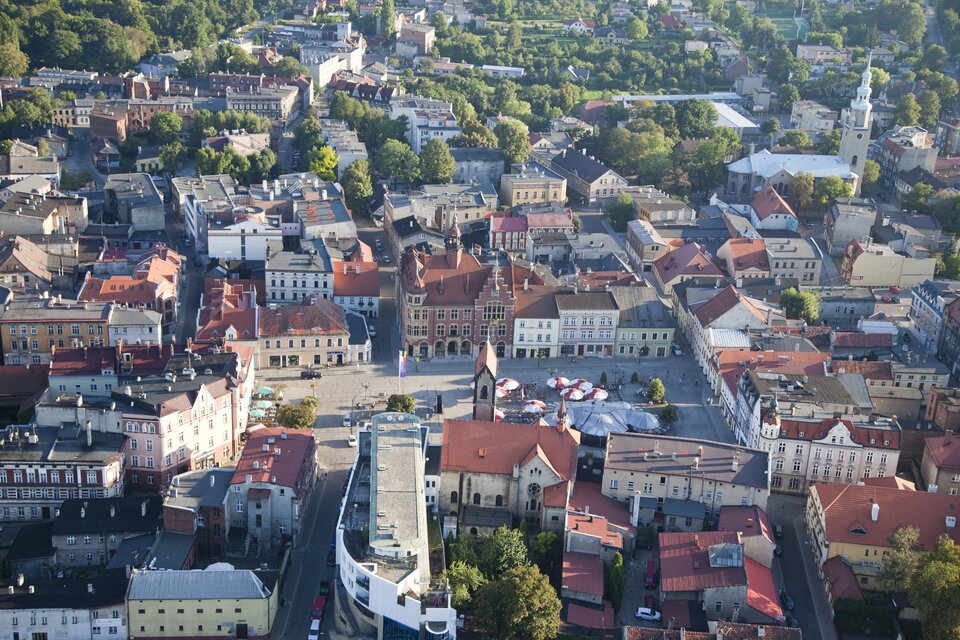Na zdjęciu przedstawiony jest fragment Tarnowskich Gór z lotu ptaka. W centrum fotografii znajduje się rynek, dookoła którego są kamienice. Od rynku odchodzi siatka ulic, a po prawej stronie widoczny jest kościół z wysoką wieżą.