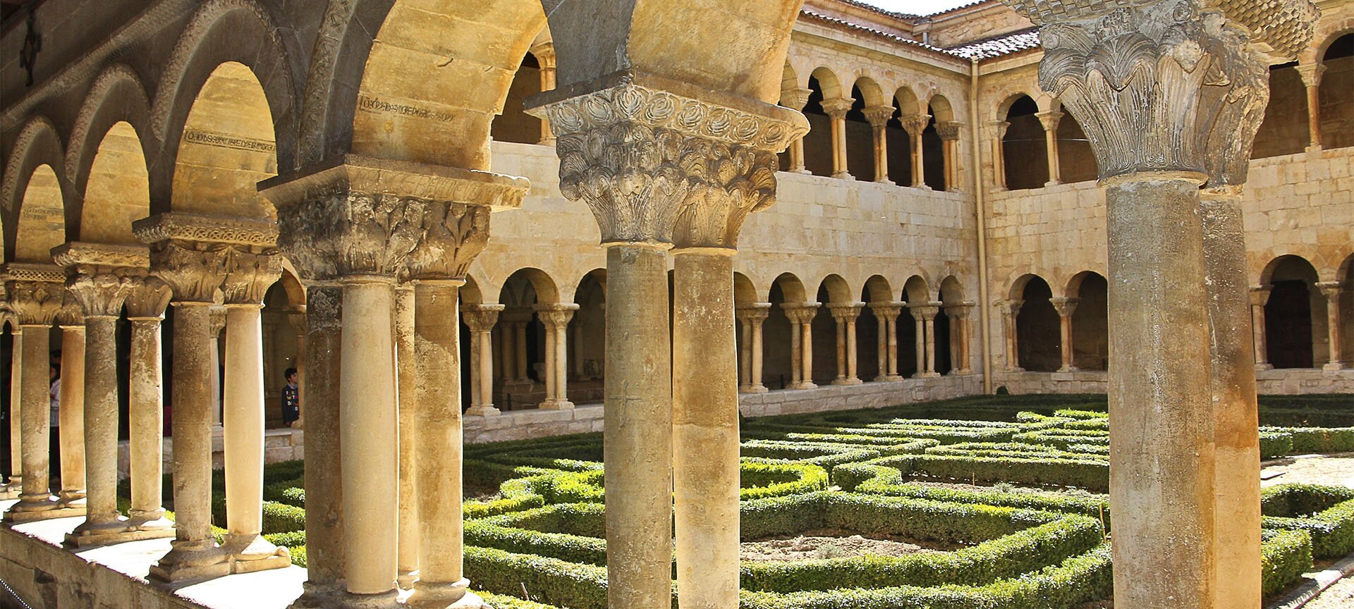 Fotografia przedstawia dziedziniec przed którym stoi kolumnada podtrzymująca loggię budynku. Budynek, jak i kolumny są zbudowane z kamienia, ktory ma kolor złotego piasku. Głowice na kolumnach mają kształt ściętego ostrosłupa, którego ściany zostały bogato przyozdobione wymodelowanymi elementami w kształcie liści akantu. Głowice opierają się na dwóch lub na czterech kolumnach. Na dziedzińcu rosną żywopłoty, które tworzą labirynt.