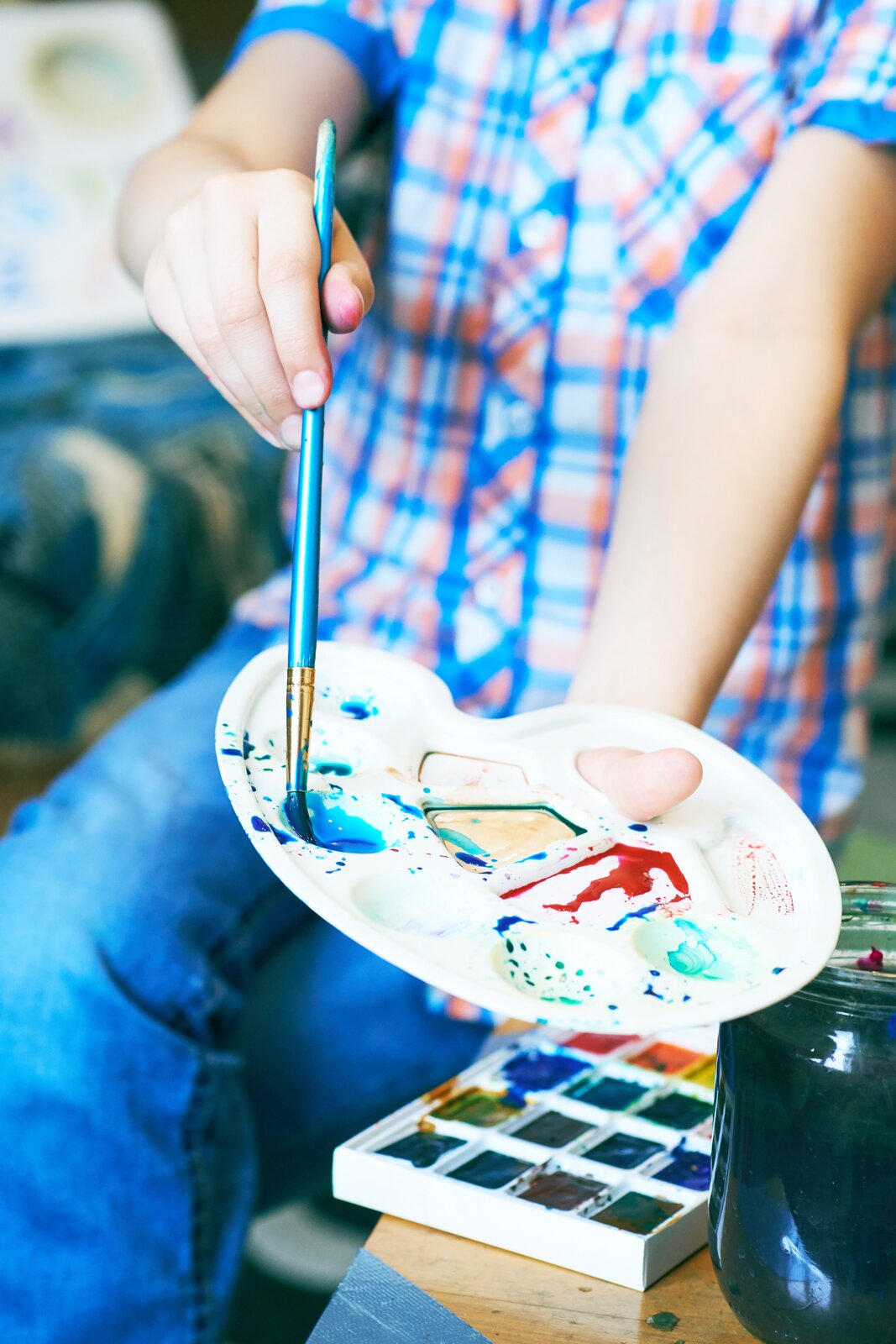 Plastikowa paletka, sposób trzymania palety Fotografia przedstawia widok na plastikową paletą trzymaną przez dziecko. Paleta została wykonana z białego materiału i ma kształt owalu, z dziurą pozwalającą na wygodne trzymanie. Widoczne są na niej farby - czerwona, żółta, zielona oraz niebieska, w którą chłopiec wkłada pędzel trzymany przez niego w prawej dłoni. Kciuk jest włożony w dziurę palety, zaś pozostała jej część opiera się na dłoni chłopca. Obok chłopca trzymającego paletę widoczny jest słoik z wodą oraz farby w zestawie składającym się z małych prostokątów. Chłopiec ma na sobie ciemne jeansy oraz granatowo‑czerwoną koszulę z krótkim rękawem w kratę. Tło jest rozmyte.