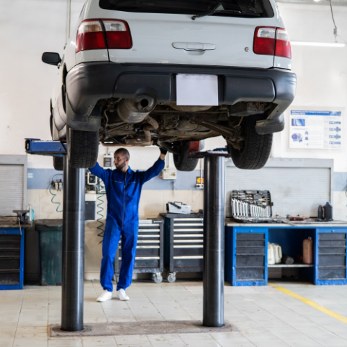 Na grafice pokazane przykładowe narzędzie warsztatowe, czyli podnośnik samochodowy. Na podnośniku znajduje się samochód, który podniesiony jest kilka metrów nad ziemią. Przed pojazdem stoi mechanik trzymający ręce na elemencie podnośnika. Ubrany jest w niebieski kombinezon.