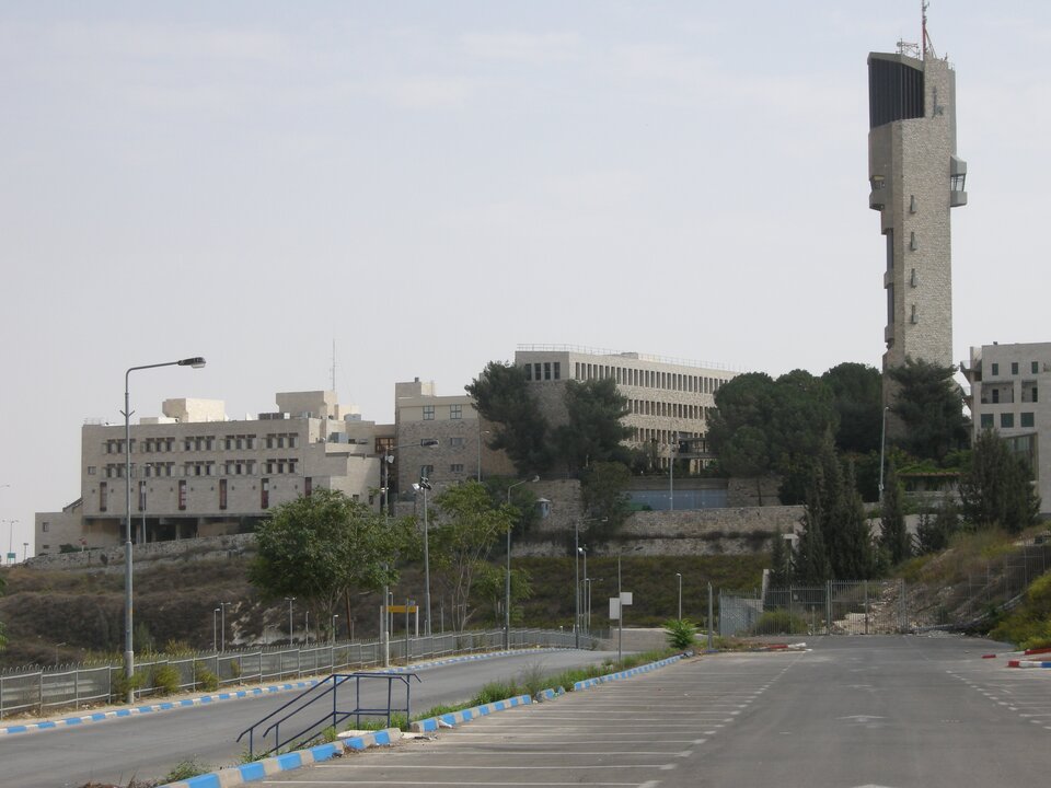 Zdjęcie przedstawia budynki Akademii Sztuk Pięknych i Wzornictwa Besaleela na górze Skopus. Budynki mają prosty, nowoczesny kształt. Nad nimi góruje wysoka, prostopadłościenna wieża. Na pierwszym planie pusty parking. 