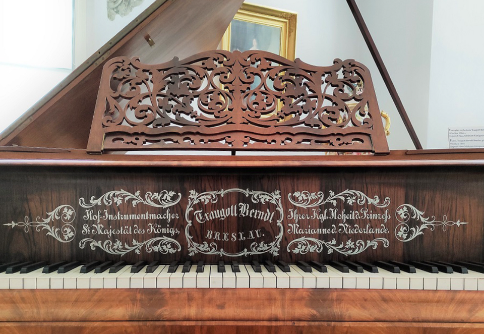 Zdjęcie przedstawia klawiaturę fortepianu, powyżej na obudowie ozdobne napisy i rzeźbiona podpórka pod nuty.
