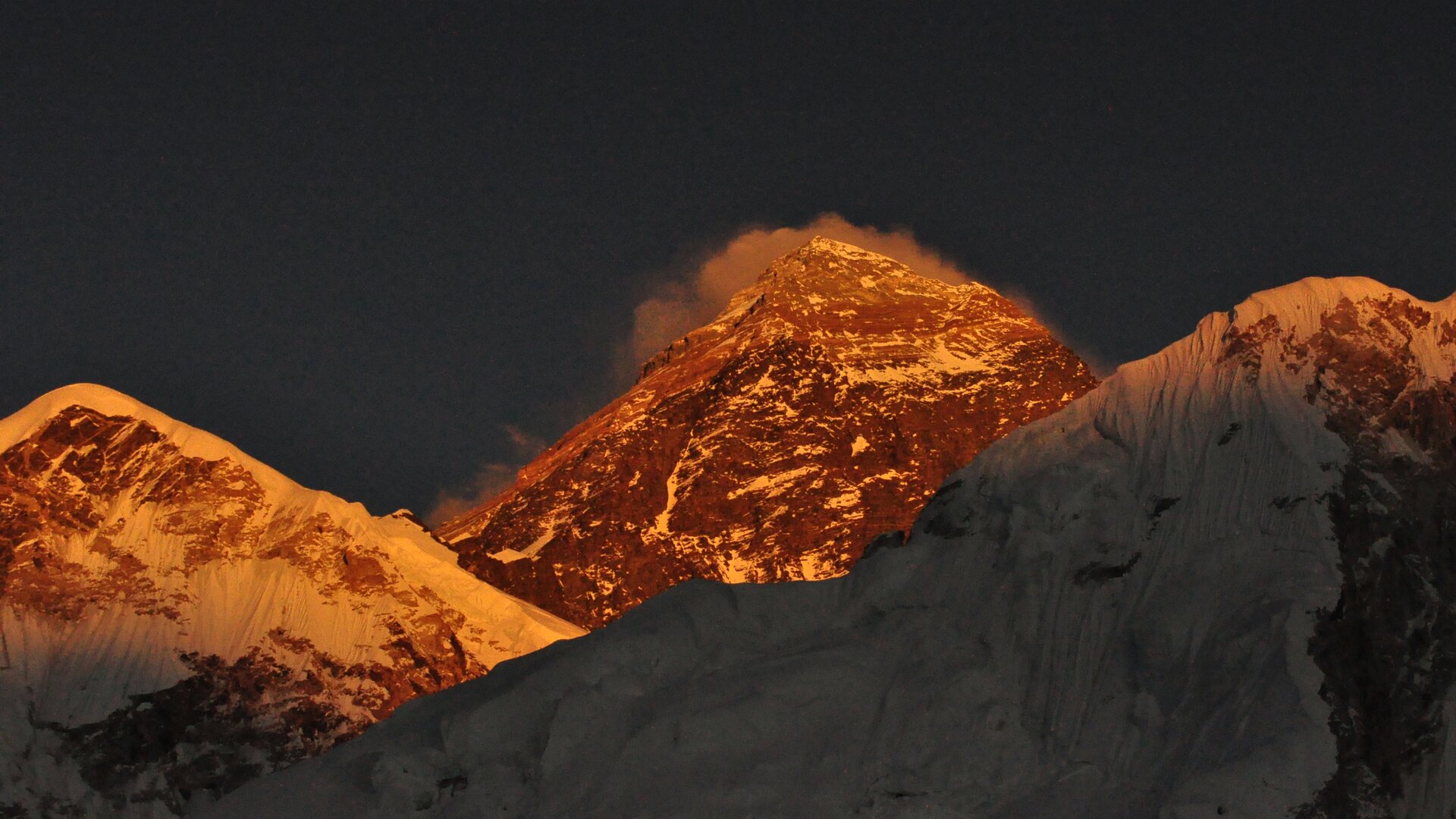 Zdjęcie przedstawia trzy szczyty oświetlone słońcem i częściowo ośnieżone. Środkowy szczyt jest najwyższy. Spowija go obłok.