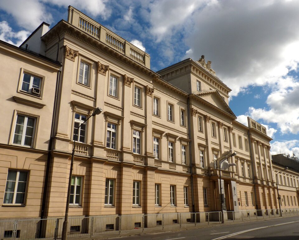 Na zdjęciu widać długi, jasnobeżowy budynek o kilku kondygnacjach, ustawiony wzdłuż ulicy. Elewacja jest symetryczna, z regularnie rozmieszczonymi prostokątnymi oknami w białych obramowaniach. Na wyższych kondygnacjach widoczne są dekoracyjne pilastry i gzymsy, a na dachu fragmenty balustrad. W środkowej części budynku znajduje się lekko wysunięty fragment z trójkątnym naczółkiem i rzeźbami na górze. Przy fasadzie stoją latarnie uliczne oraz metalowe barierki oddzielające chodnik od jezdni. Na pierwszym planie widoczna jest asfaltowa ulica. W tle znajduje się niebo z jasnymi chmurami.