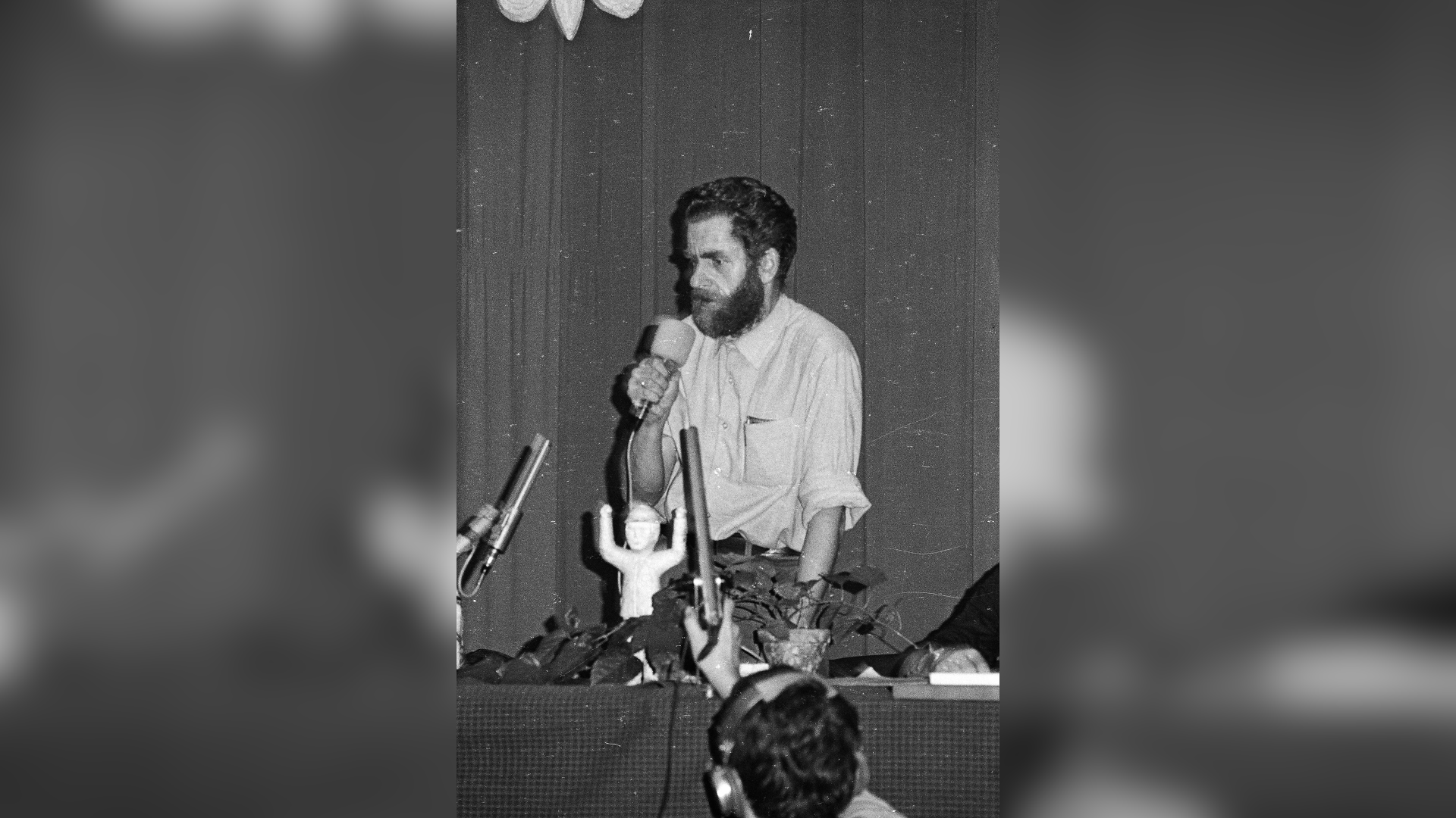 Fotografia przedstawiająca mężczyznę w sile wieku. Postać stoi przed stołem, na którym ustawiono mikrofony i doniczkę z kwiatem. Mężczyzna przedstawiony jest w całości, ukazuje lewy profil. Mężczyzna ma owalną twarz, zmarszczone czoło, gęste brwi, głęboko osadzone oczy, duży nos oraz obfity zarost na twarzy. Włosy ma gęste, lekko falujące. Ubrany jest w koszulę z rękawami podwiniętymi do wysokości łokci. Spodnie zapięte skórzanym paskiem. W prawej ręce trzyma mikrofon. Na serdecznym palcu ma obrączkę. Lewą rękę opiera o stół. W tle wiszące zasłony. 