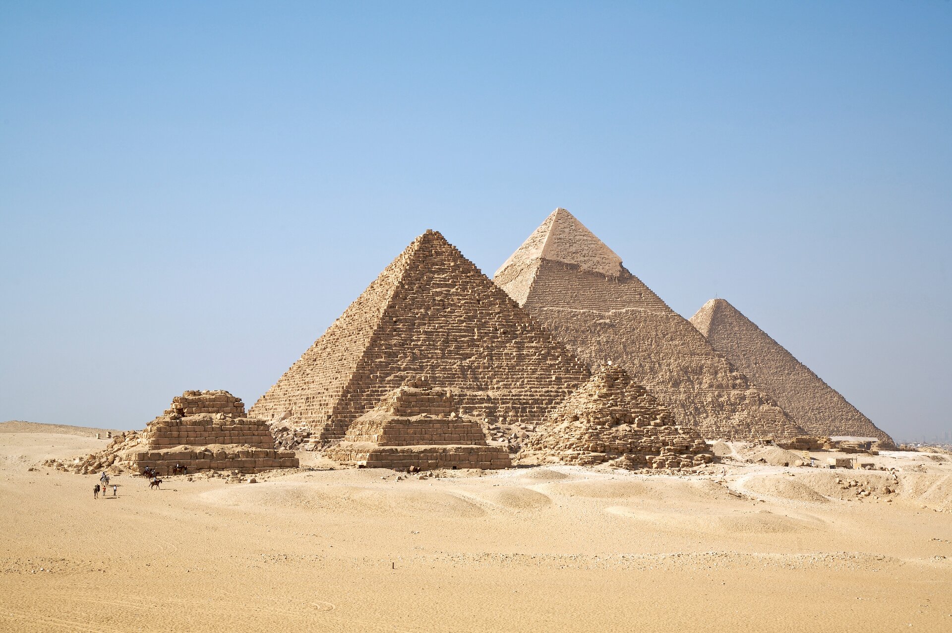 Zdjęcie przedstawia piramidy w Gizie na tle nieba. Wokół piramid przechadzają się ludzie.