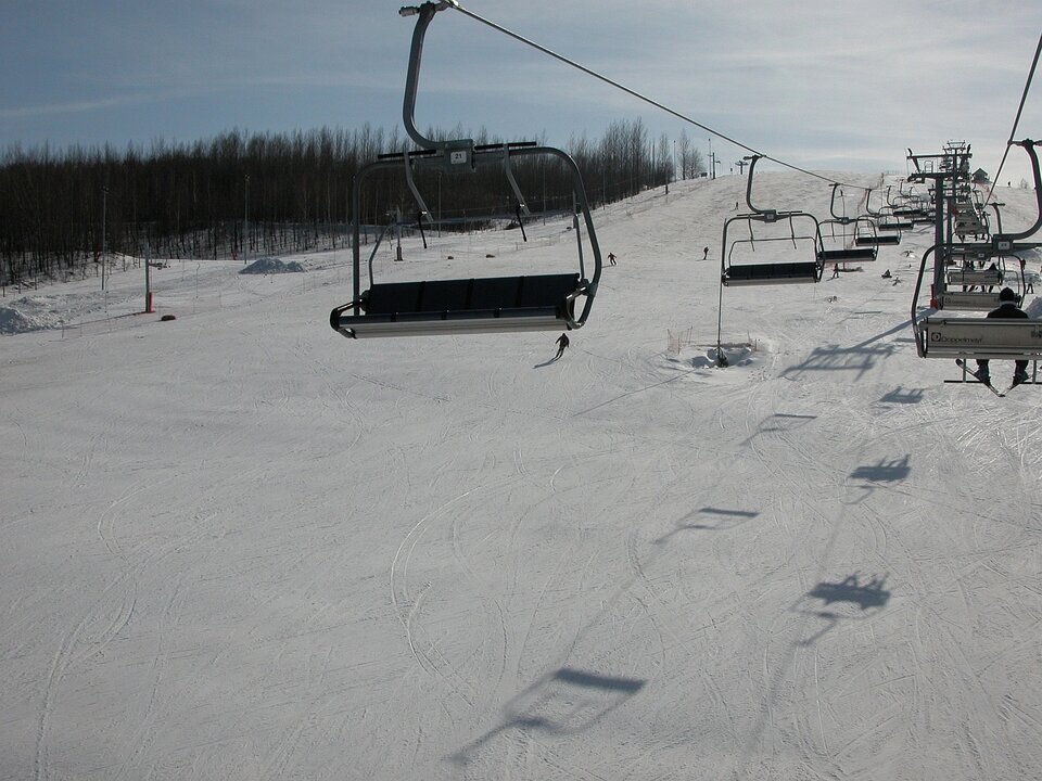 Zdjęcie przedstawia stok narciarski, na którym leży śnieg i na którym znajduje się kilku narciarzy. Nad stokiem jest wyciąg krzesełkowy. Stok jest dosyć łagodny.  