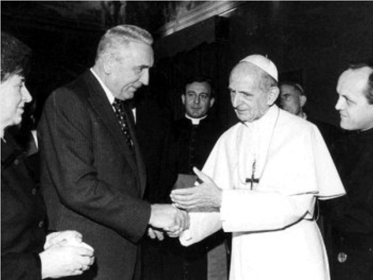 Zdjęcie przedstawia witających się Edwarda Gierka i papieża Pawła VI. Mężczyźni ściskają sobie prawe dłonie. Wokół nich zgromadzeni są ludzie.