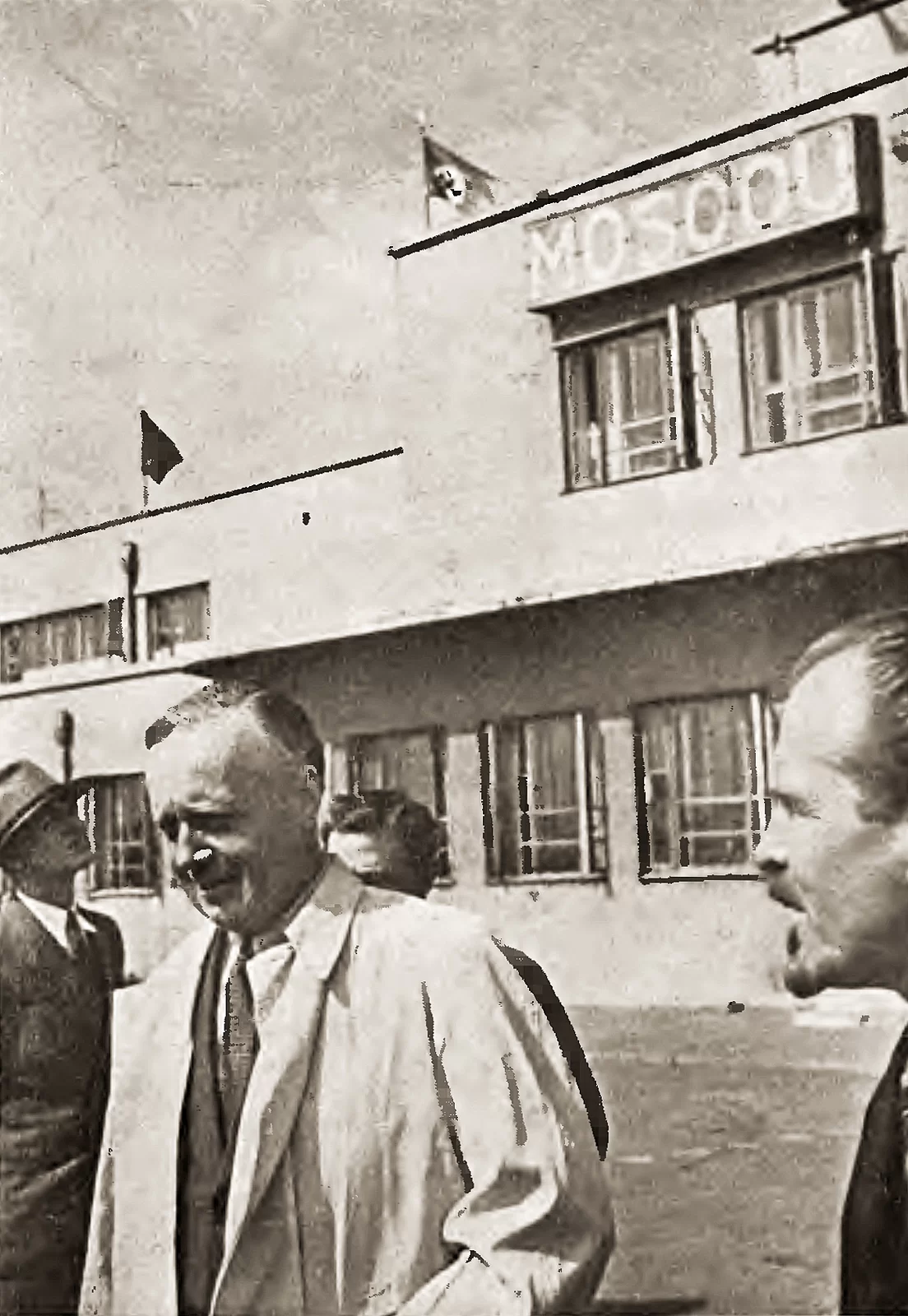 Zdjęcie przedstawia Ribbentropa stojącego przed budynkiem lotniska. Ma krótkie, zaczesane na bok włosy, ubrany jest w garnitur z jasnym płaszczem. Towarzyszy mu trzech mężczyzn w garniturach. Na budynku za nim powiewają flagi ZSRS i Trzeciej Rzeszy. Na fasadzie z oknami znajduje się napis Moskwa.
