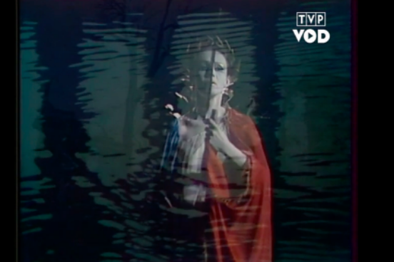 Fotografia przedstawia nakładające się na siebie dwa obrazy. Pośrodku stoi kobieta w sile wieku. Postać ukazuje w całości owalną twarz. Kobieta ma gładkie czoło, jasne brwi, zwężone oczy, prosty nos i wąskie usta. Jej głowę zdobi wieniec. Kobieta oburącz trzyma włócznię. Okryta jest czerwoną pelerynę. Drugie zdjęcie przedstawia powierzchnię wody. Utworzyły się na niej okręgi.