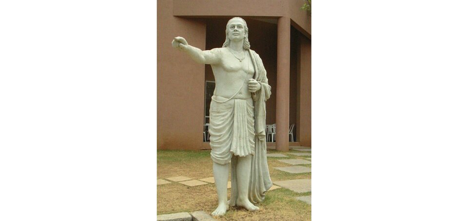 Zdjęcie przedstawia rzeźbę mężczyzny o włosach do ramion. Postać ma podniesioną na wysokość barków prawą rękę i pokazuje palcem wskazującym w przestrzeń. Jego głowa jest lekko uniesiona do góry. Mężczyzna ma nagi tors, jest przepasany materiałem, ma zawieszoną pelerynę na lewym barku, jest boso. Mężczyzna ma dwa wisiorki.