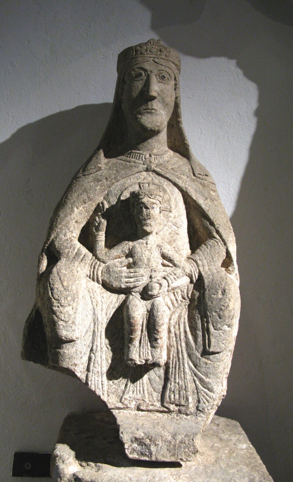 Fotografia przedstawia kamienną figurę Tronującej Madonny z Goźlic. Rzeźba ma uproszczoną formę i kształt. Przedstawia siedzącą na tronie Maryję z Dzieciątkiem Jezus, które trzyma z przodu kolanach i przytrzymuje rękami. Maryja na głowie posiada chustę spływającą do tyłu, jej wyraz twarzy jest surowy. Ubrana jest w długą szatę. Dzieciątko Jezus nad głową posiada aureolę. 