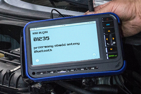 Grafika przedstawia tester diagnostyczny. Jest to niewielkie urządzenie podobne do tabletu z wyświetlaczem i przyciskami. Na jego ekranie widoczny jest kod błędu B1 dwa trzy pięć, a pod nim napis: przerwany obwód anteny bluetooth.