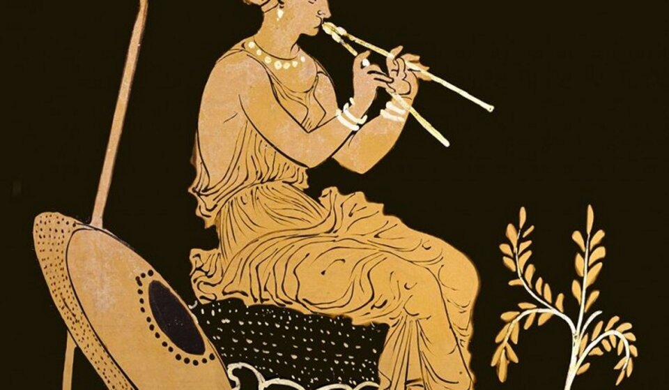 Ilustracja przedstawia siedzącą kobietę, która gra na instrumencie. Kobieta w policzkach trzyma powietrze. Ubrana jest w szatę, obok niech stoi tarcza. Instrument muzyczny widoczny na ilustracji to aulos - stanowiły go jedna lub dwie niezależne, cylindryczne piszczałki wyposażone w podwójny stroik.