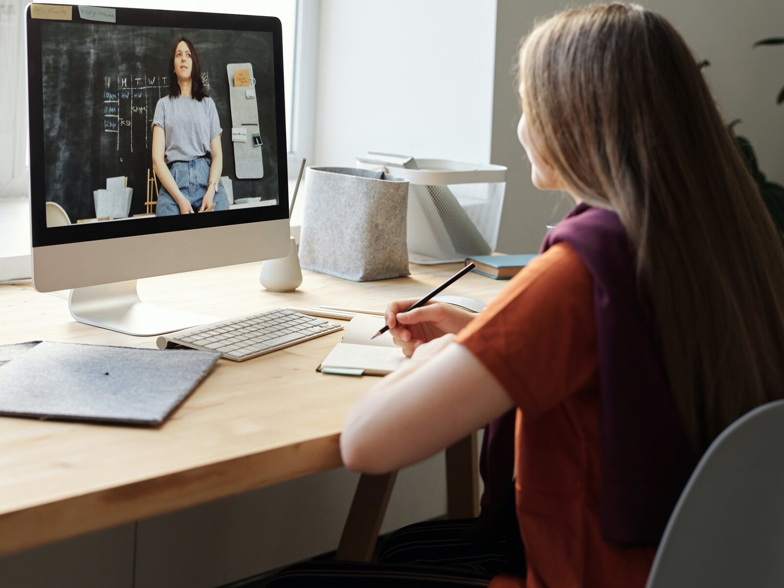 Zdjęcie przedstawia nastolatkę siedzącą przy biurku, w ujęciu z lewego boku. Nastolatka ma długie jasne włosy, jest ubrana w pomarańczową koszulkę z krótkim rękawem i kamizelkę. Przed nią na biurku widoczny jest otwarty notes, nad którym w prawej dłoni trzyma ołówek. Na wprost na biurku stoi monitor komputera, klawiatura, pojemnik na ołówki, pojemnik na dokumenty. Na monitorze komputera widoczna jest młoda kobieta siedząca na tle czarnej szkolnej tablicy z napisami.