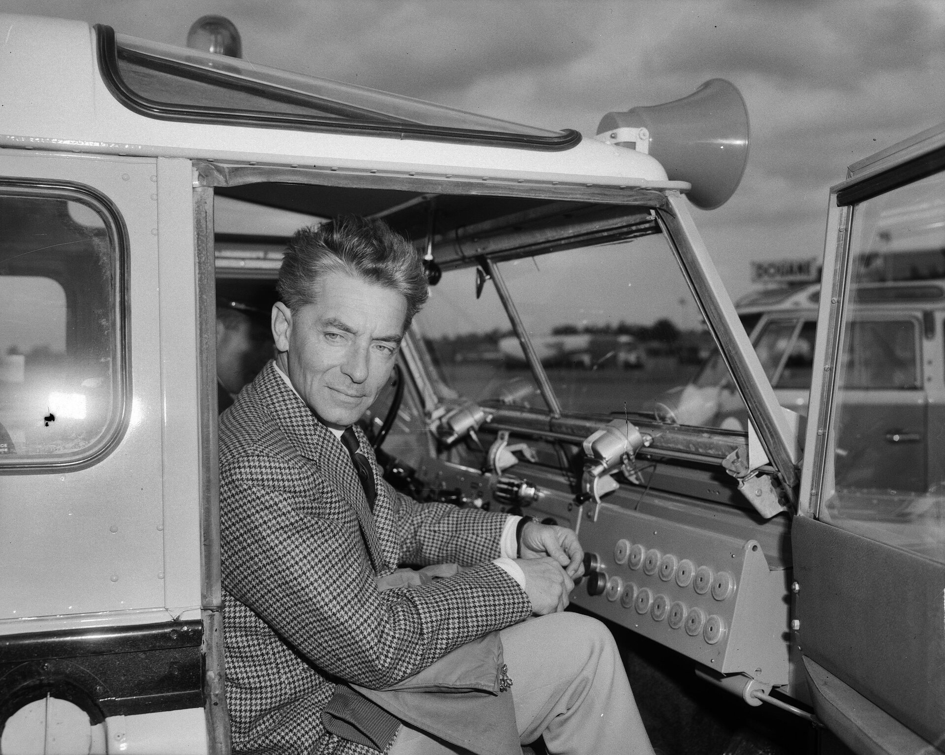 Zdjęcie przedstawiające Herberta von Karajana, fotografia Joop van Bilsen. Czarno‑biała fotografia ukazuje postać mężczyzny, siedzącego w samochodzie. Drzwi samochodu są otwarte. Mężczyzna ubrany jest w marynarkę w kratę. 