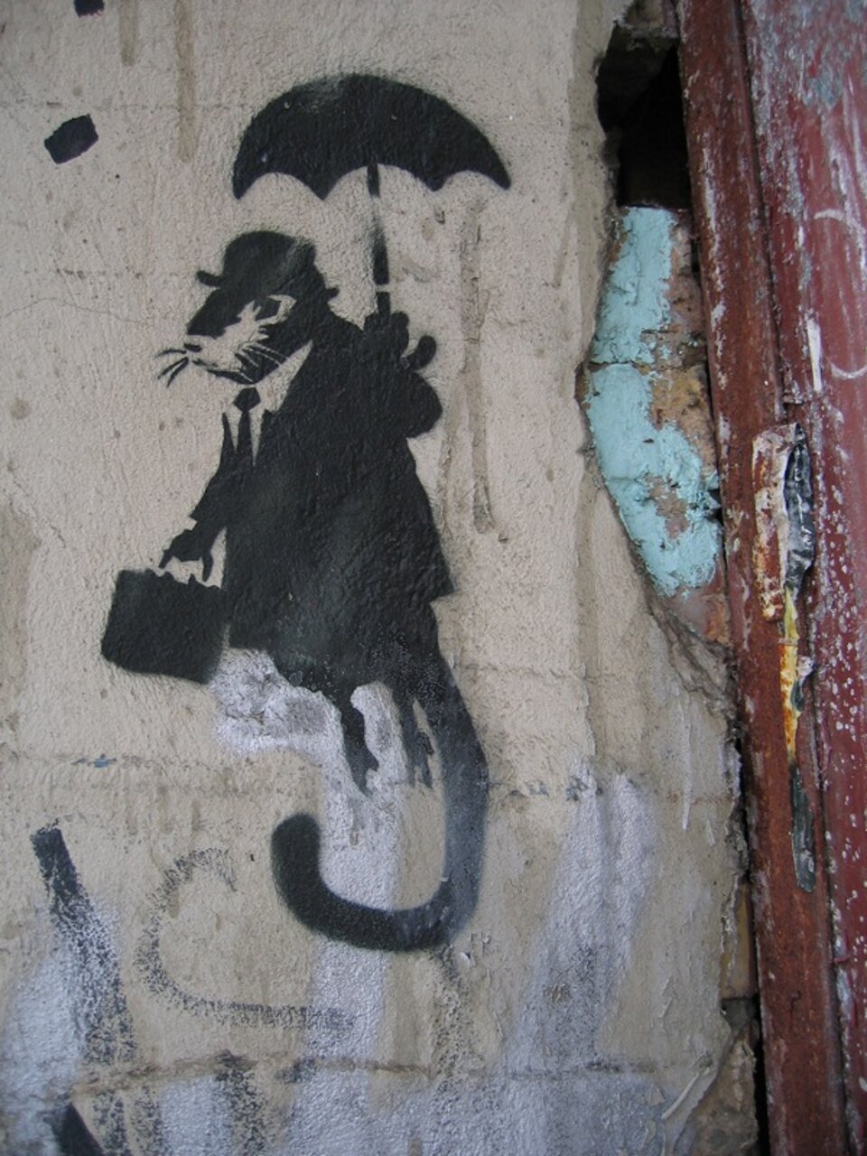 Ilustracja przedstawia mural prezentujący szczura w białej koszuli, z zawiązanym krawatem oraz w płaszczu i kapeluszu, który idąc na dwóch łapach w pozostałych dwóch trzyma w jednej teczkę a w drugiej parasol. Spod płaszcza wystaje jego długi ogon. Mural wykonany jest w czarnym kolorze na szarym tle. 