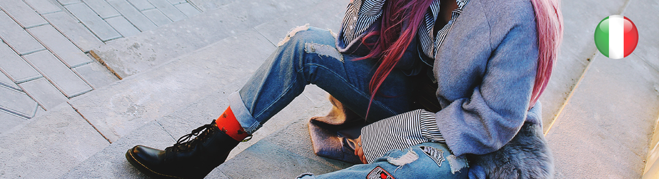 Zdjęcie zrobione z góry przedstawia osobę siedząca na betonowych schodach. Niewidoczna jest twarz tej osoby. Ubrana jest w buty typu glany, podarte dżinsy,  koszulę w paski, płaszcz i szal.