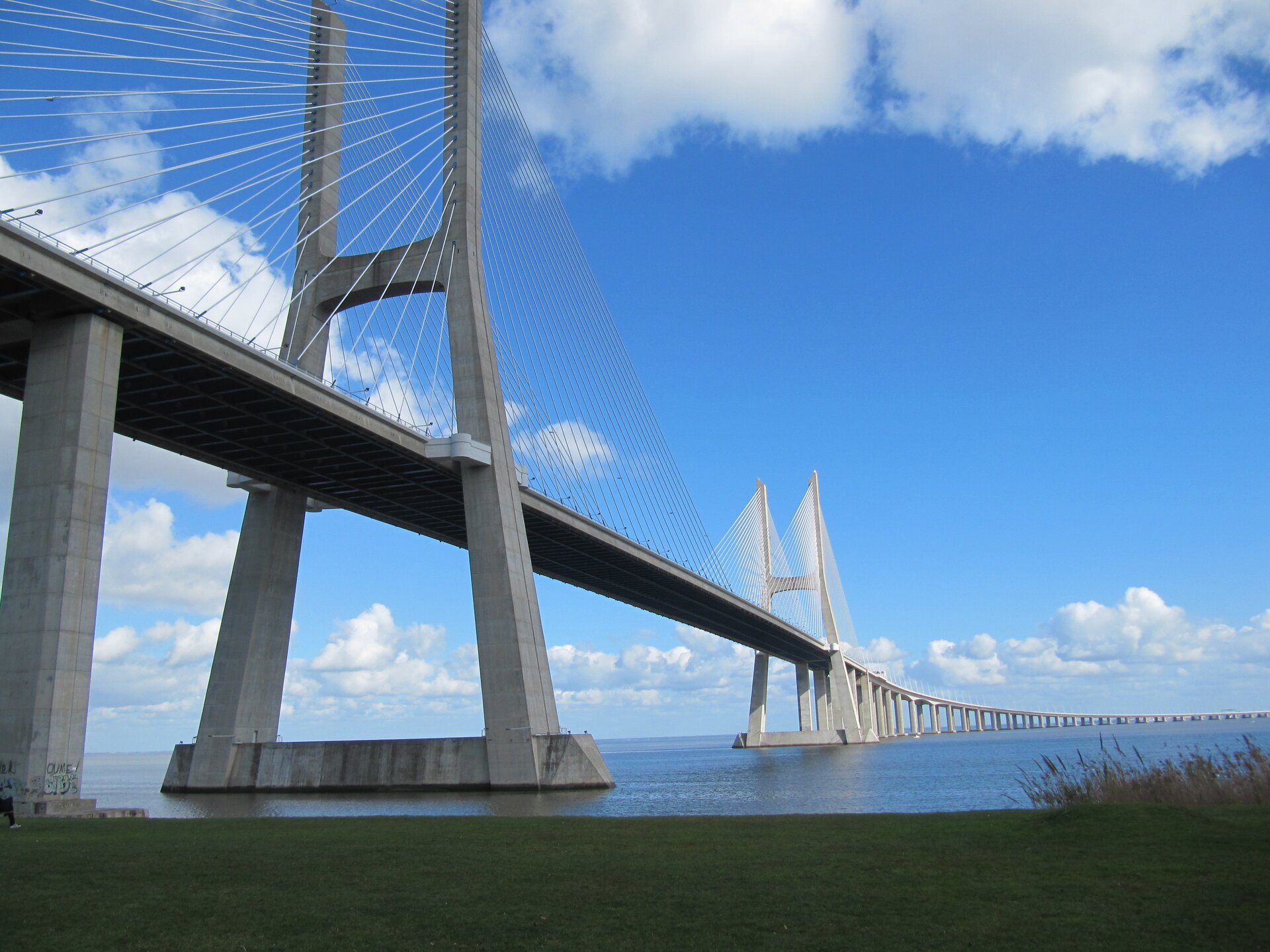 Zdjęcie przedstawia most Vasco da Gama  w Lizbonie, który jest spektakularnym przykładem wykorzystania betonu we współczesnym budownictwie. Estetyka mostu jest nowoczesna i prosta. Jest to konstrukcja wisząca o długości około siedemnastu i dwóch dziesiątych kilometra, przecinająca rzekę Tag. Jego charakterystycznym elementem są dwa wysokie filary w kształcie żagli, które podtrzymują konstrukcję wiszącą. Most wykorzystuje konstrukcję wiszącą, gdzie pionowe liny podtrzymują most przez zastosowanie naciągu. Ta technologia pozwala na znaczne zwiększenie rozpiętości mostu, jednocześnie minimalizując obciążenie filarów. Most Vasco da Gama  pełni rolę zarówno drogową, jak i kolejową. Posiada dwie oddzielne jezdnie dla ruchu samochodowego oraz torowisko dla pociągów.