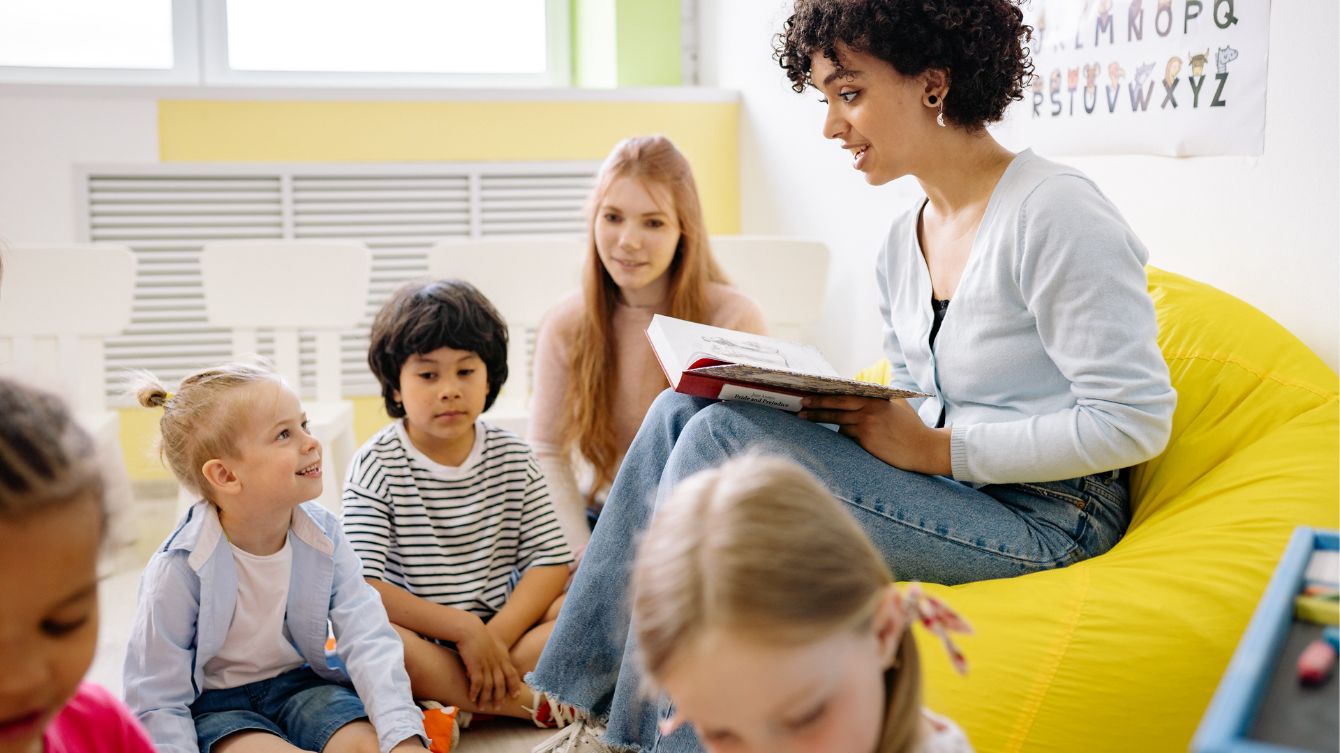 Zdjęcie przedstawia dzieci siedzące na podłodze oraz młodą, uśmiechniętą kobietę z krótkimi, kręconymi włosami. Kobieta siedzi w fotelu z otwartą książką na kolanach. Dzieci siedzą na podłodze przed nią. Widoczne są dwie dziewczynki oraz jeden chłopiec. Na pierwszym planie widoczne są fragmenty głów dwóch dziewczynek, które siedzą tyłem do kobiety w fotelu i zajmują się czymś innym.