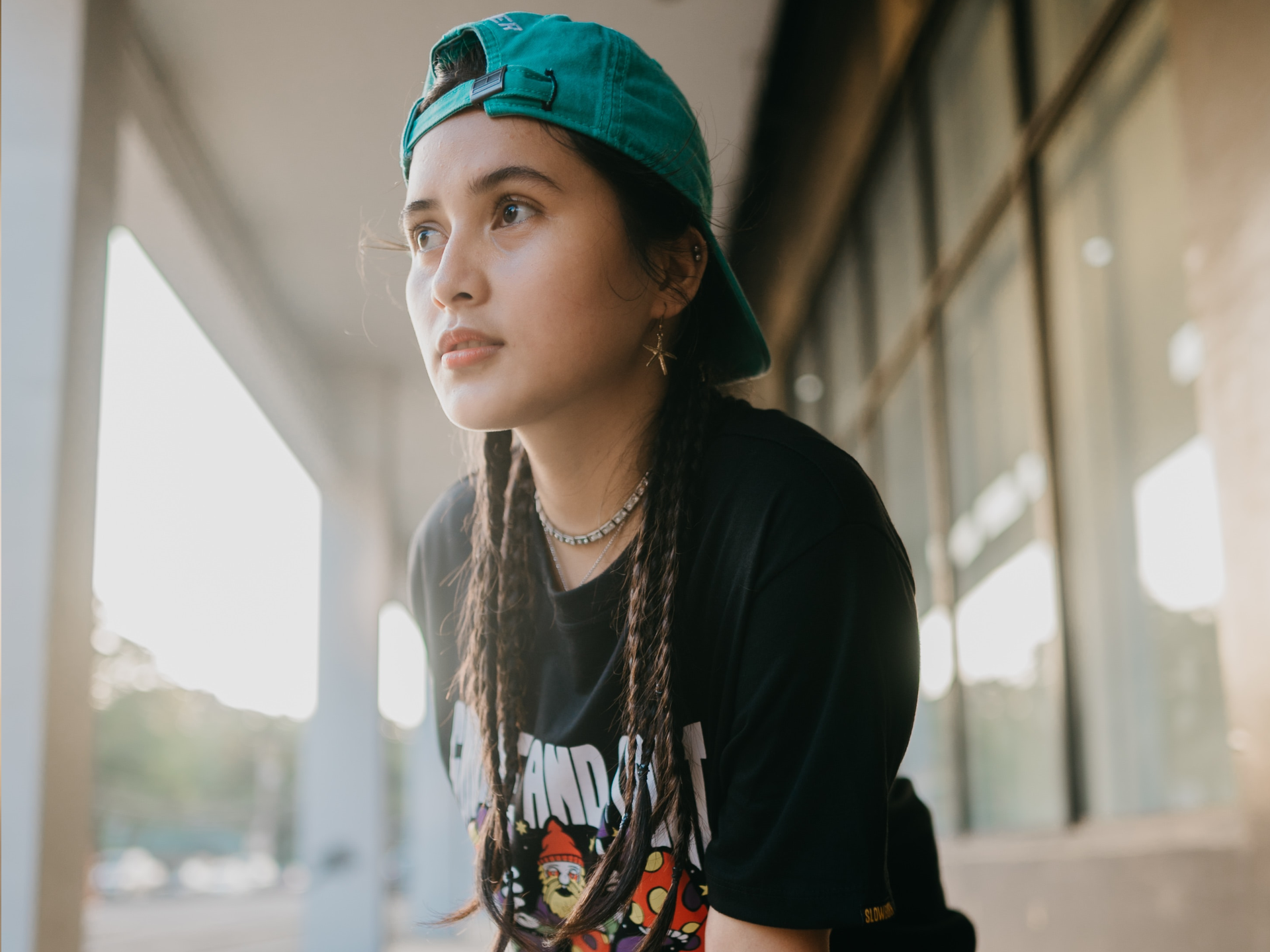 Zdjęcie przedstawia młodą dziewczynę, w ujęciu od pasa w górę, zwróconą w lewo. Dziewczyna ma długie, ciemne włosy, zaplecione w warkoczyki, a na głowie czapkę odwróconą daszkiem do tyłu. Jest ubrana w czarną koszulę z krótkim rękawem i rysunkiem z przodu. W lewym uchu dziewczyny widoczny jest złoty kolczyk w kształcie gwiazdy oraz dwa małe kolczyki w kształcie kulek. Na szyi widoczny jest srebrny naszyjnik i łańcuszek.