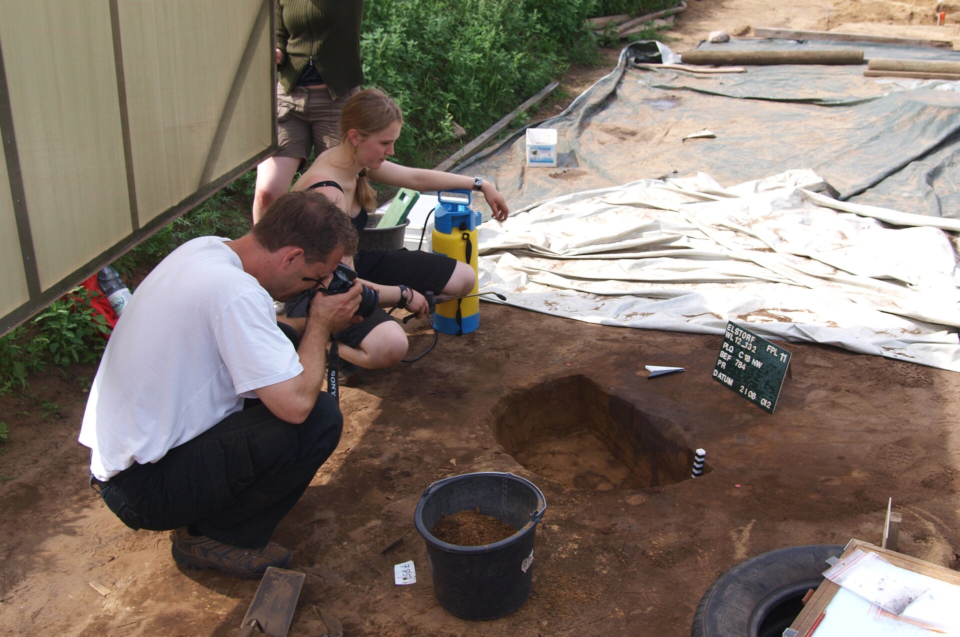 Zdjęcie przedstawia kobietę i mężczyznę kucających nad dołem i fotografujących znaleziska w nim odkryte.