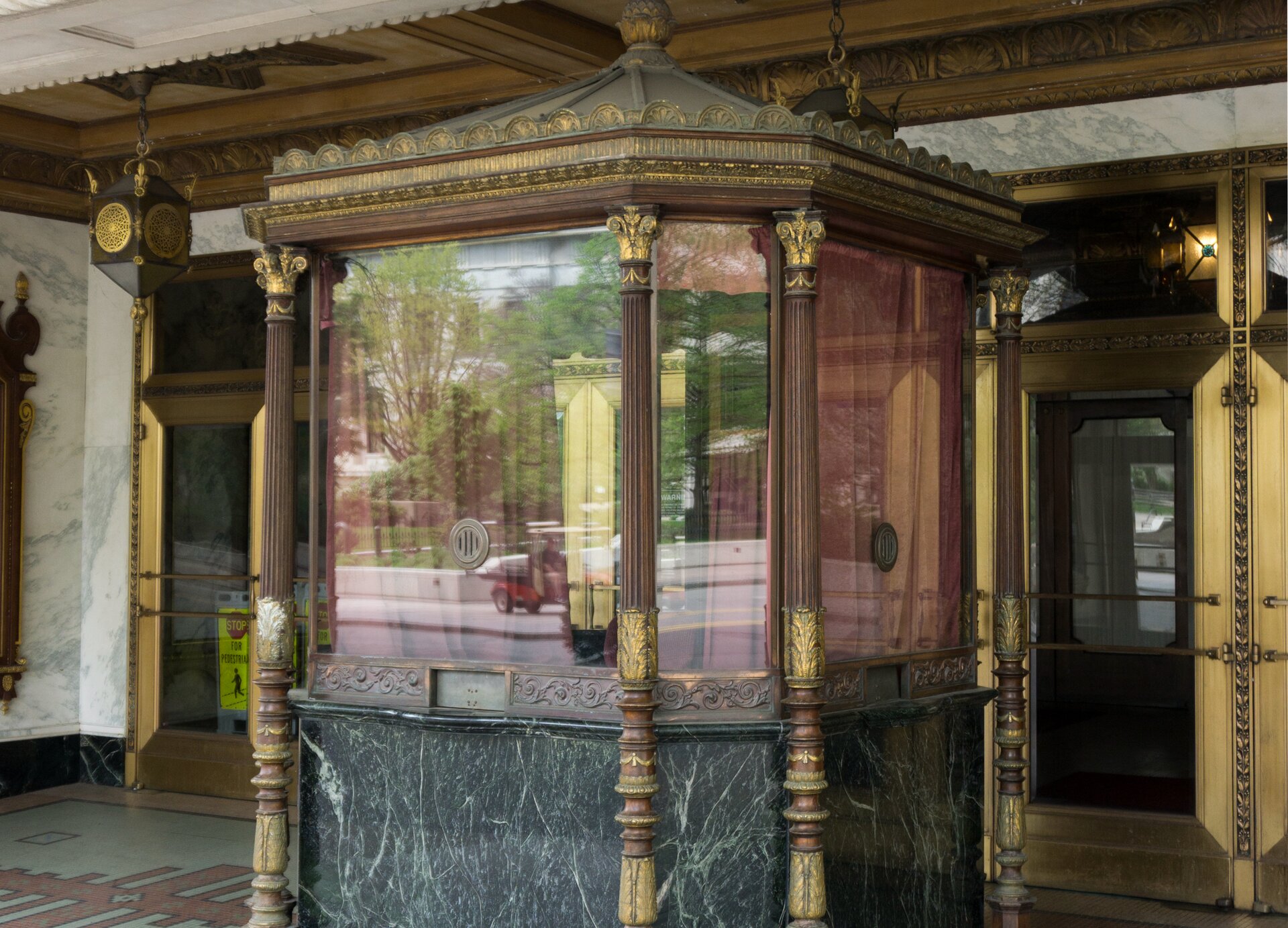 Zdjęcie przedstawia wejście do zabytkowego budynku  teatru z widoczną pośrodku zamkniętą kasą biletową. Widoczny jest niewielka konstrukcja, od połowy do góry przeszklona ze wszystkich stron, której narożniki zdobione są drewnianymi kolumnami ze złoconymi kapitelami. Dolna część wyłożona jest marmurem, a na dekorowanym pasie biegnącym między marmurem a szybami widoczne są małe, zasunięte okienka kas. Całość zwieńczona jest złoconym gzymsem i schodzącym się pośrodku dachem zakończonym złoconym zdobieniem. Witryna zasunięta jest brązowymi kotarami, a w środku widoczne jest krzesło. Za kasą widoczne są wejścia do teatru.