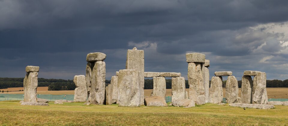 Fotografia przedstawia Stonehenge w Anglii. Widok ukazuje panoramiczny widok na kamienny krąg. Budowla składa się z dużych pionowych bloków kamiennych (megalitów), niektóre z nich połączone są poziomymi belkami kamiennymi, tworząc charakterystyczne konstrukcje składające się z  dwóch pionowych kamieni i ułożonego na nich poziomego głazu. Bloki mają chropowatą powierzchnię i nieregularne krawędzie. Trawa wokół konstrukcji jest przystrzyżona, pożółkła. W tle widoczna jest otwarta, zielona przestrzeń z lasem na horyzoncie, a niebo jest ciemnoniebieskie i zachmurzone. 