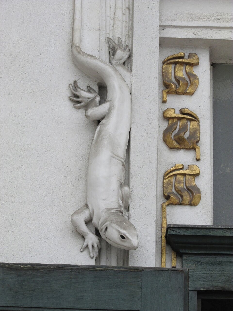 Ilustracja przedstawia detal widoczny na elewacji Pawilonu Secesji Wiedeńskiej. Detal przedstawia jaszczurkę, której autorem jest Josef Maria Olbrich. Jaszczurka jest koloru srebrnego i schodzi po ścianie. Obok jaszczurki widoczne są złote ornamenty.