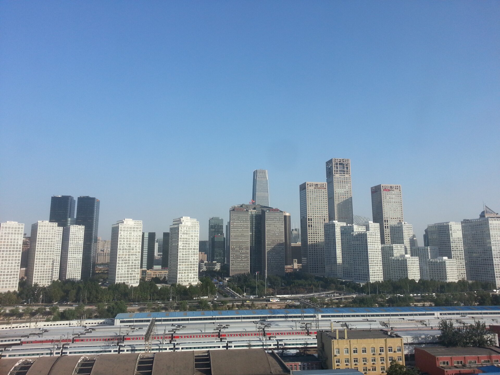 Na zdjęciu na pierwszym planie widać niską zabudowę - dachy budynków. Za nimi znajduje się szerokie torowisko z pięcioma liniami kolejowymi, na których stoją gęsto obok siebie białe wagony pasażerskie. Na drugim planie widać jezdnię wielopasmową z gęsto posadzone drzewa. Na trzecim planie, zajmującym centralne miejsce na zdjęciu widać gęstą zabudowę miejską z bardzo wysokimi wieżowcami. Przedstawione plany zajmują połowę kadru od dołu. Nad wieżowcami rozciąga się szarobłękitne niebo. Przy wieżowcach dostrzegalny jest smog.